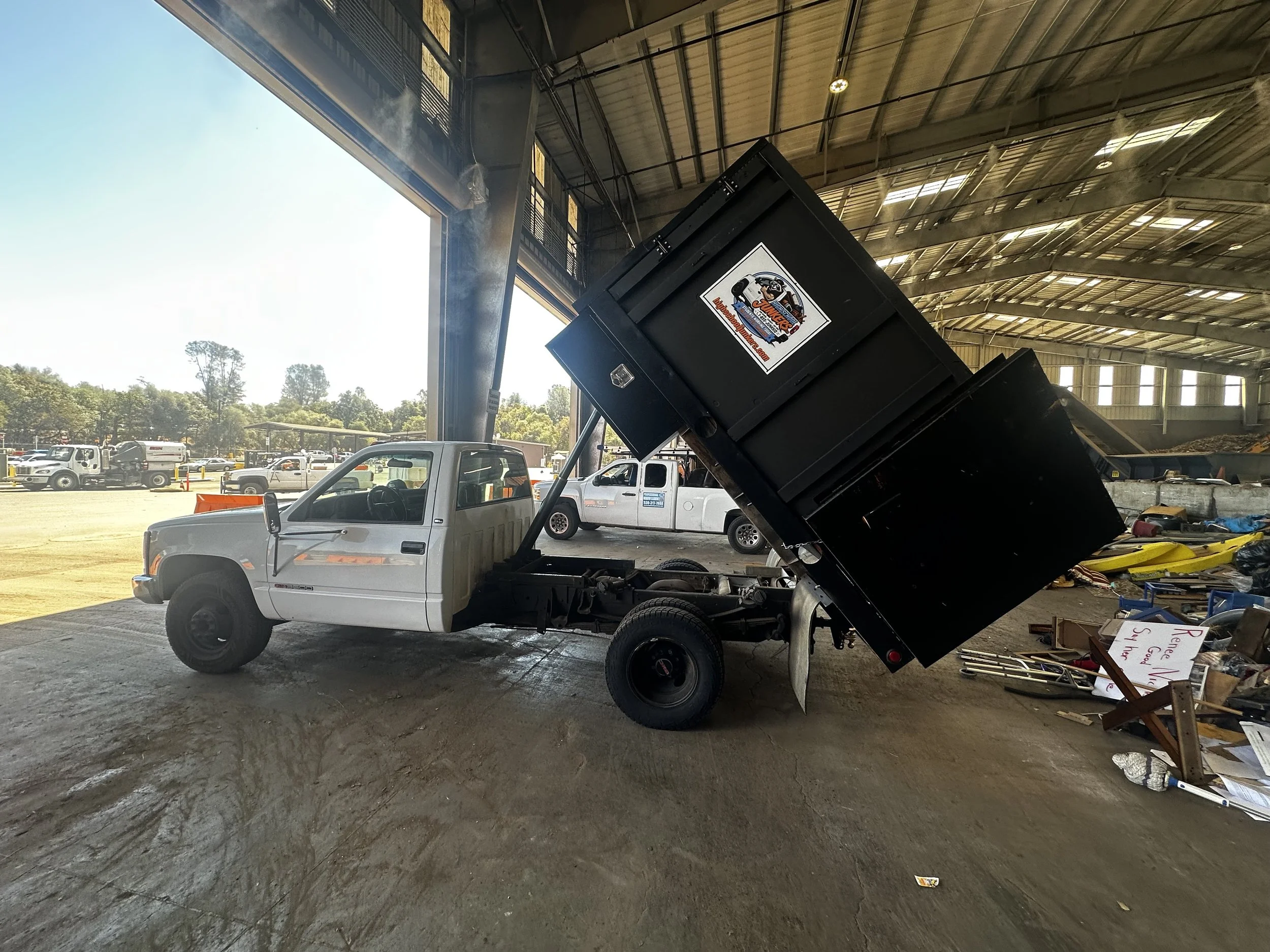 The image shows an indoor area with a lowered-ceiling metal roof and open large garage-style doors leading outside. There are multiple vehicles, including trucks and cars, parked inside and outside. In the foreground, there is a white pickup truck with a black dump trailer attached, which is raised in the air. The trailer has a sticker that reads 'Junkers' and a logo. The interior floor appears to be concrete with some scattered items, and there are signs and banners around.