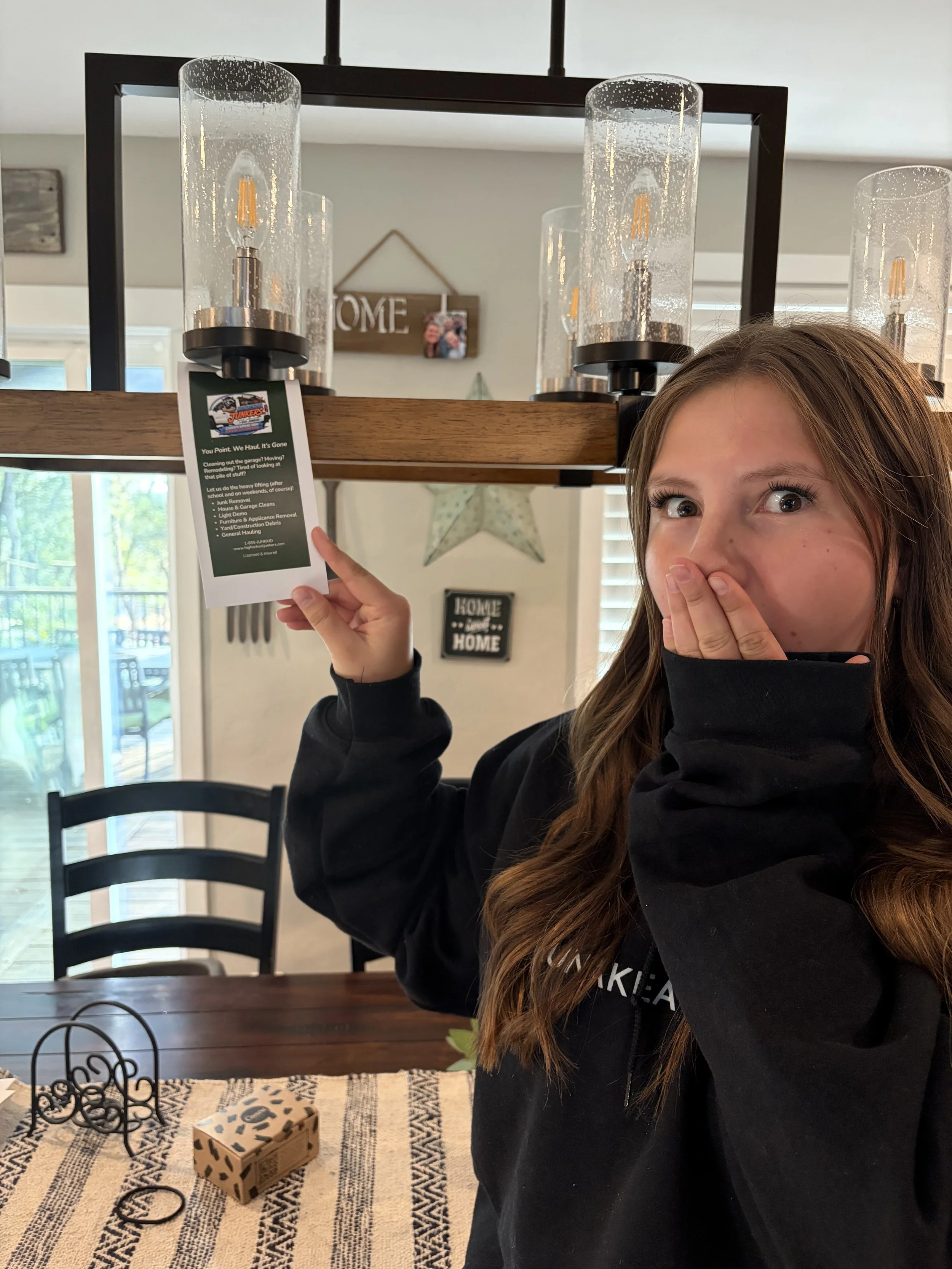 A young girl with long brown hair wearing a black hoodie covers her mouth with her hand while holding a white card with the Junkers logo, standing in a kitchen with a table and black chairs, and a wooden shelf with glass candle holders on top. There is a sign that says 'HOME' hanging on the wall in the background.