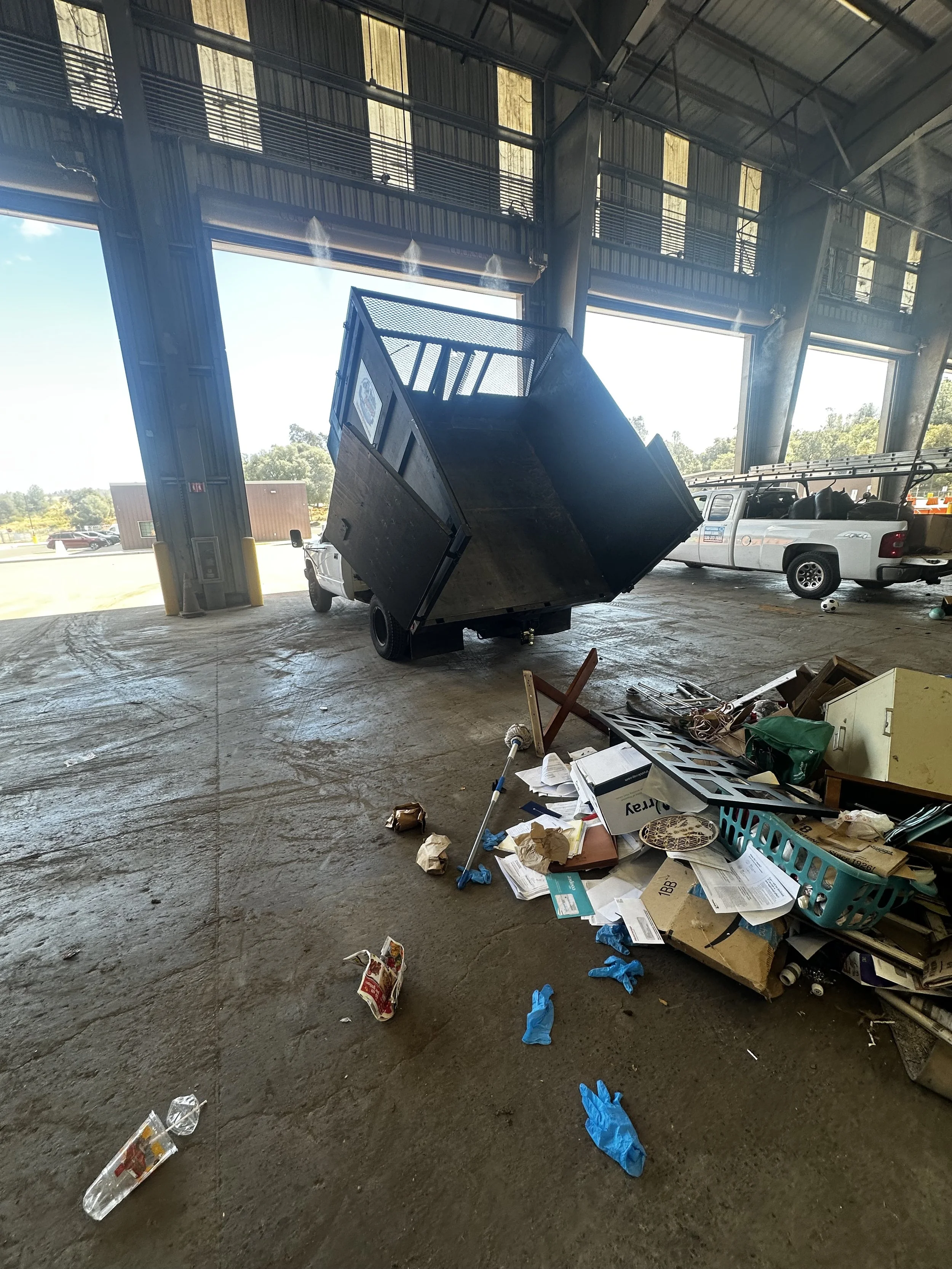 Indoor parking garage with a tilted black trailer, scattered trash, and a pickup truck nearby. Sunlight coming through open garage doors, concrete floor, and miscellaneous items strewn across the ground.