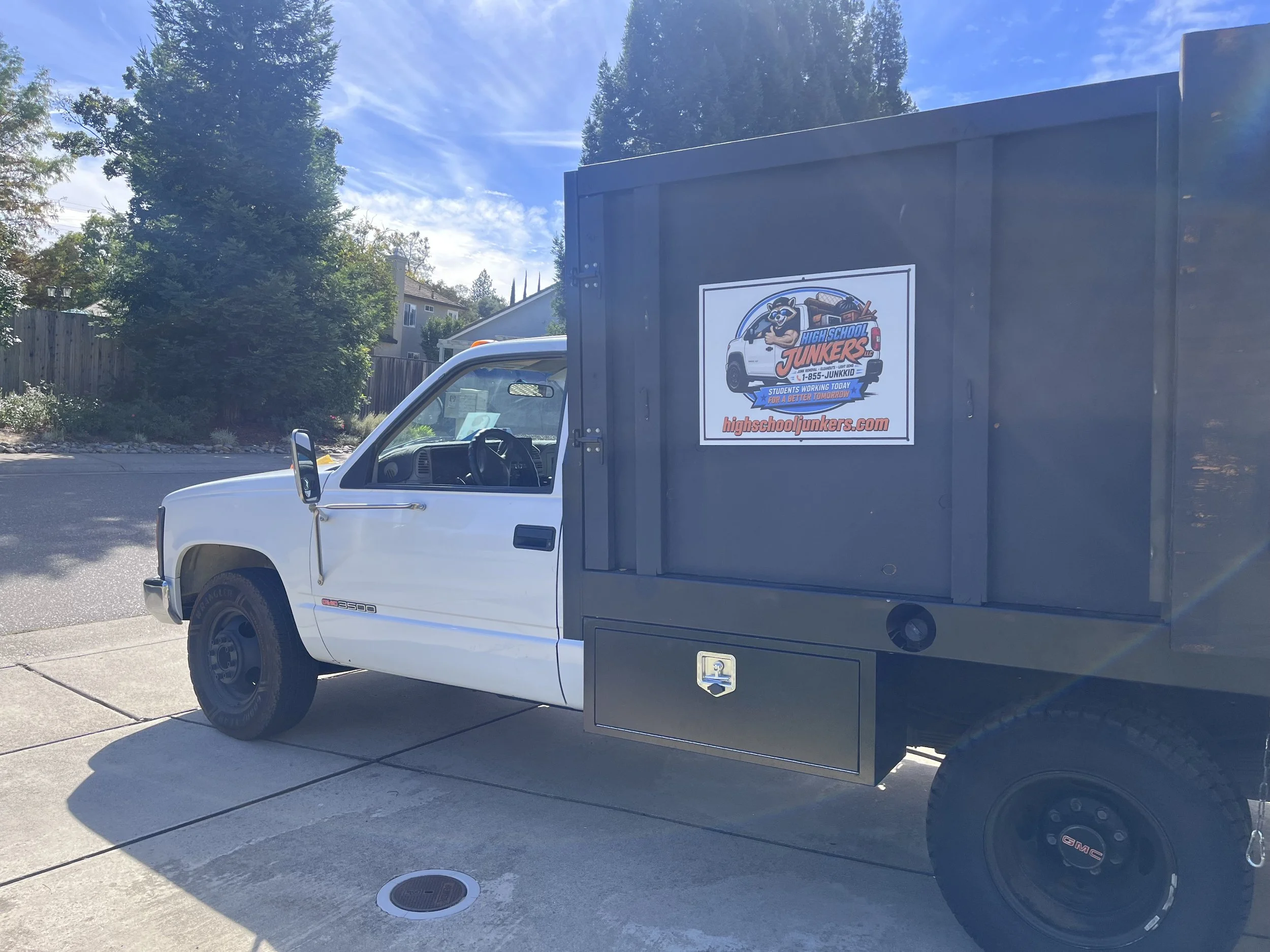 A white pickup truck with a black enclosed cargo area attached to it. The cargo area has a sign for High School Junkers, featuring a cartoon raccoon mascot, a phone number, and a website URL.