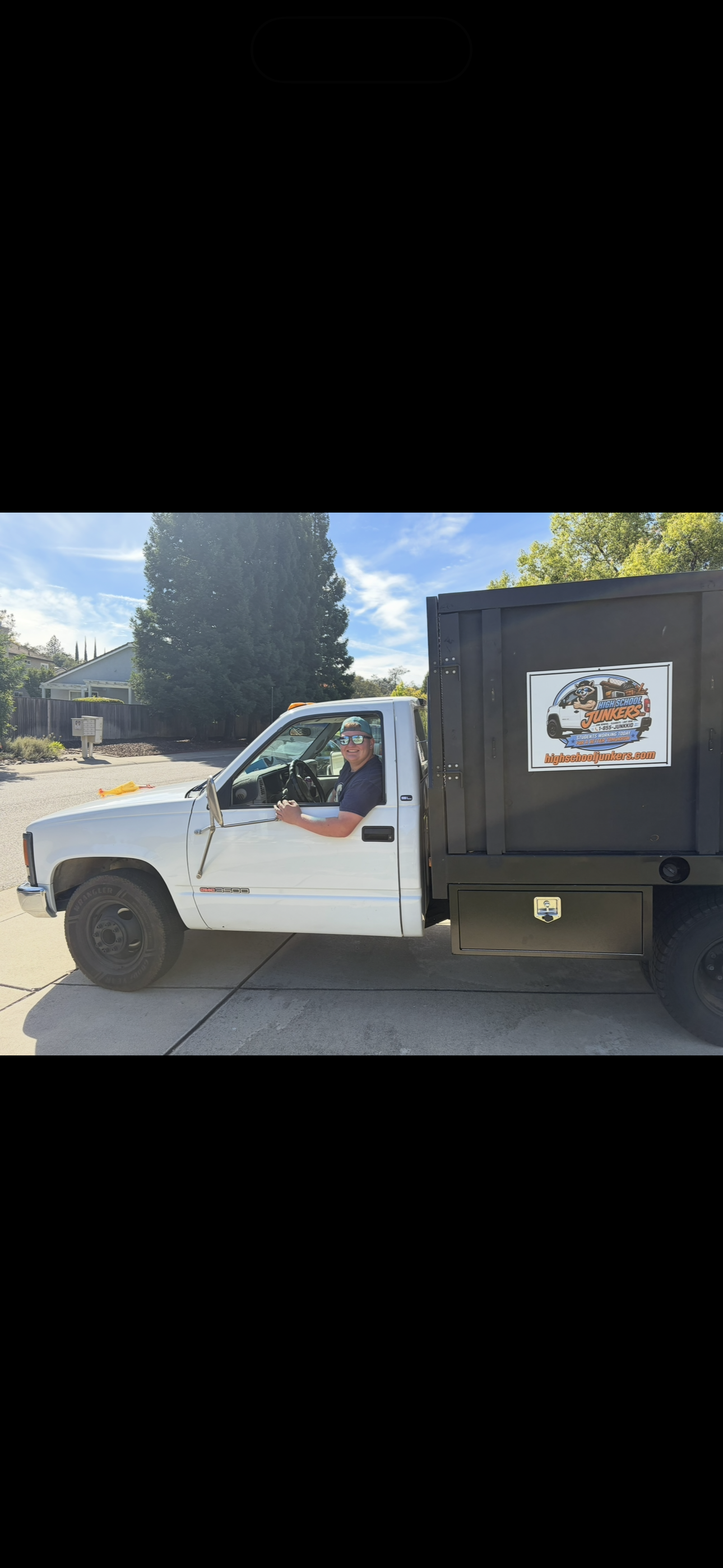 A man sitting in the driver's seat of a white pickup truck with a black storage box on the back, parked on a suburban driveway. The truck has a sign on the side that reads 'High School Junkers.' The man is smiling, wearing sunglasses and a cap, and the background shows trees, houses, and a blue sky.