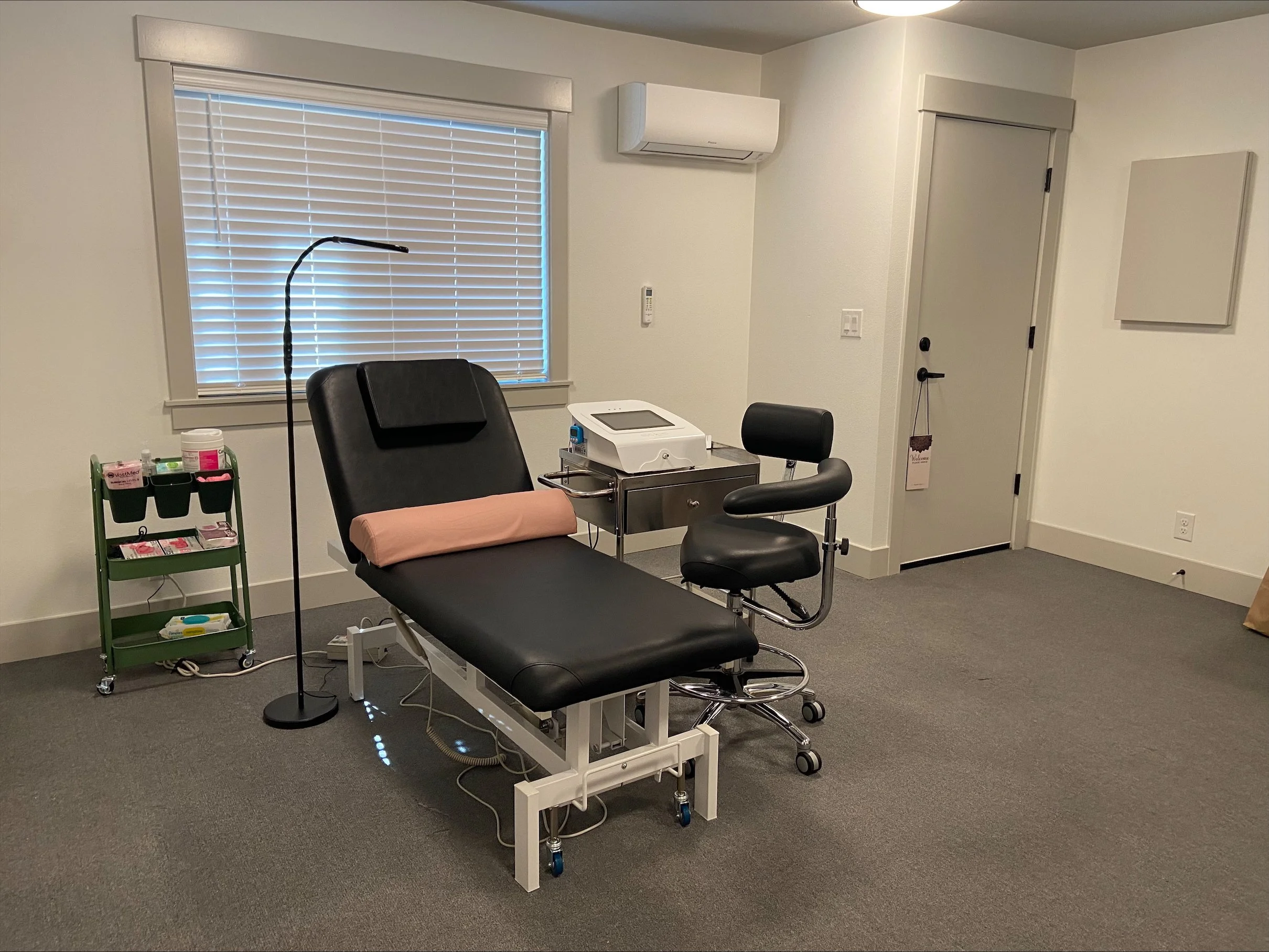 A doctor's examination room with a medical chair, a rolling cart with medical supplies, a window with blinds, a wall-mounted air conditioner, and a door with a welcome sign.