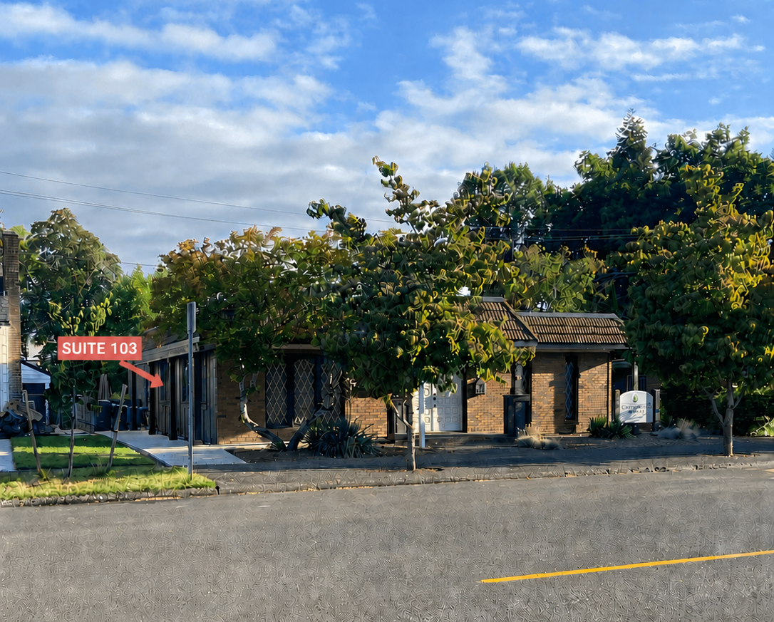 A brick building with a white door, surrounded by trees and bushes, with a sign that reads 'Suite 103' and a street view with a single yellow line.