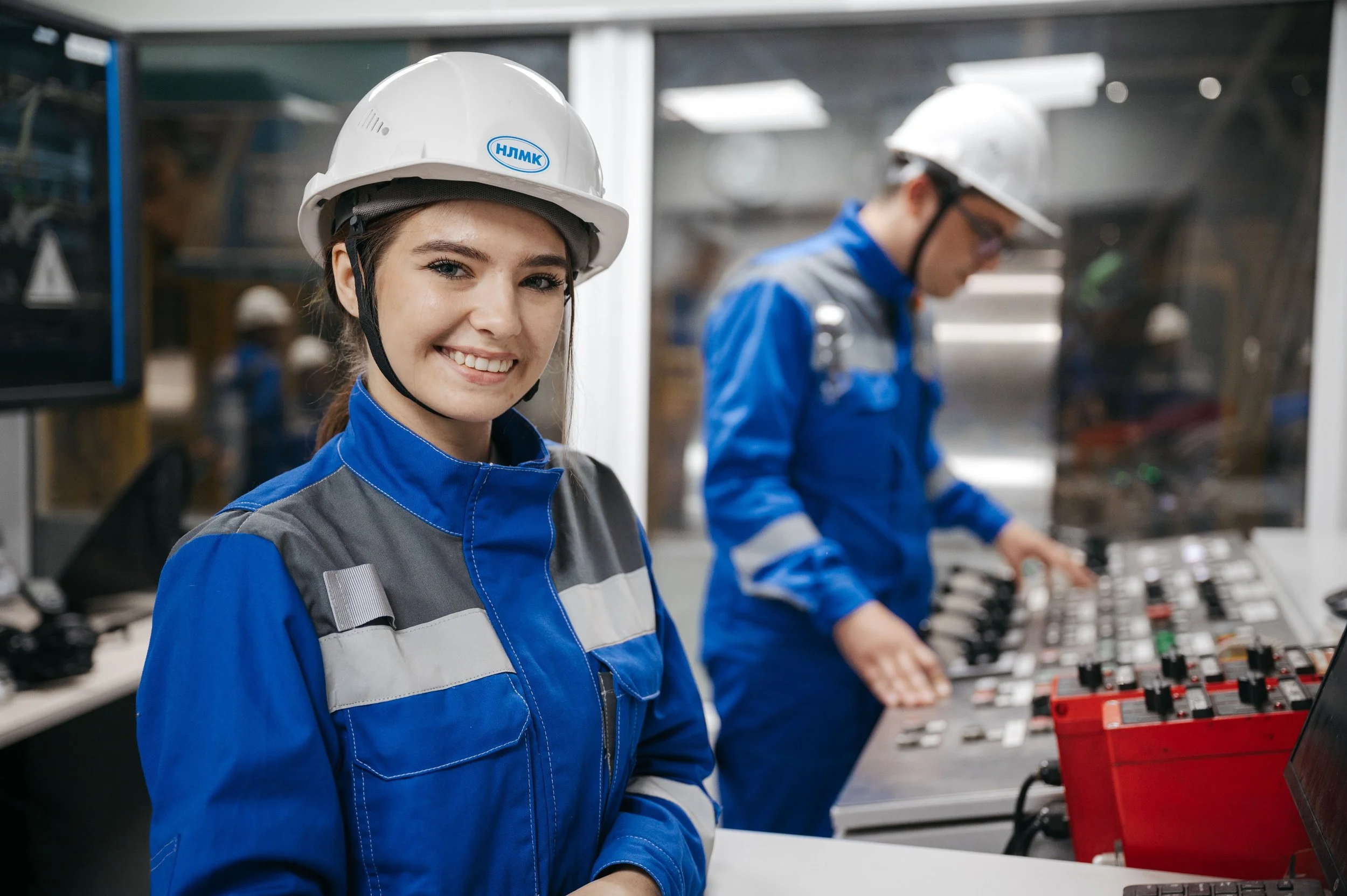Two women in blue work uniforms and white safety helmets working in a control room with electronic equipment.