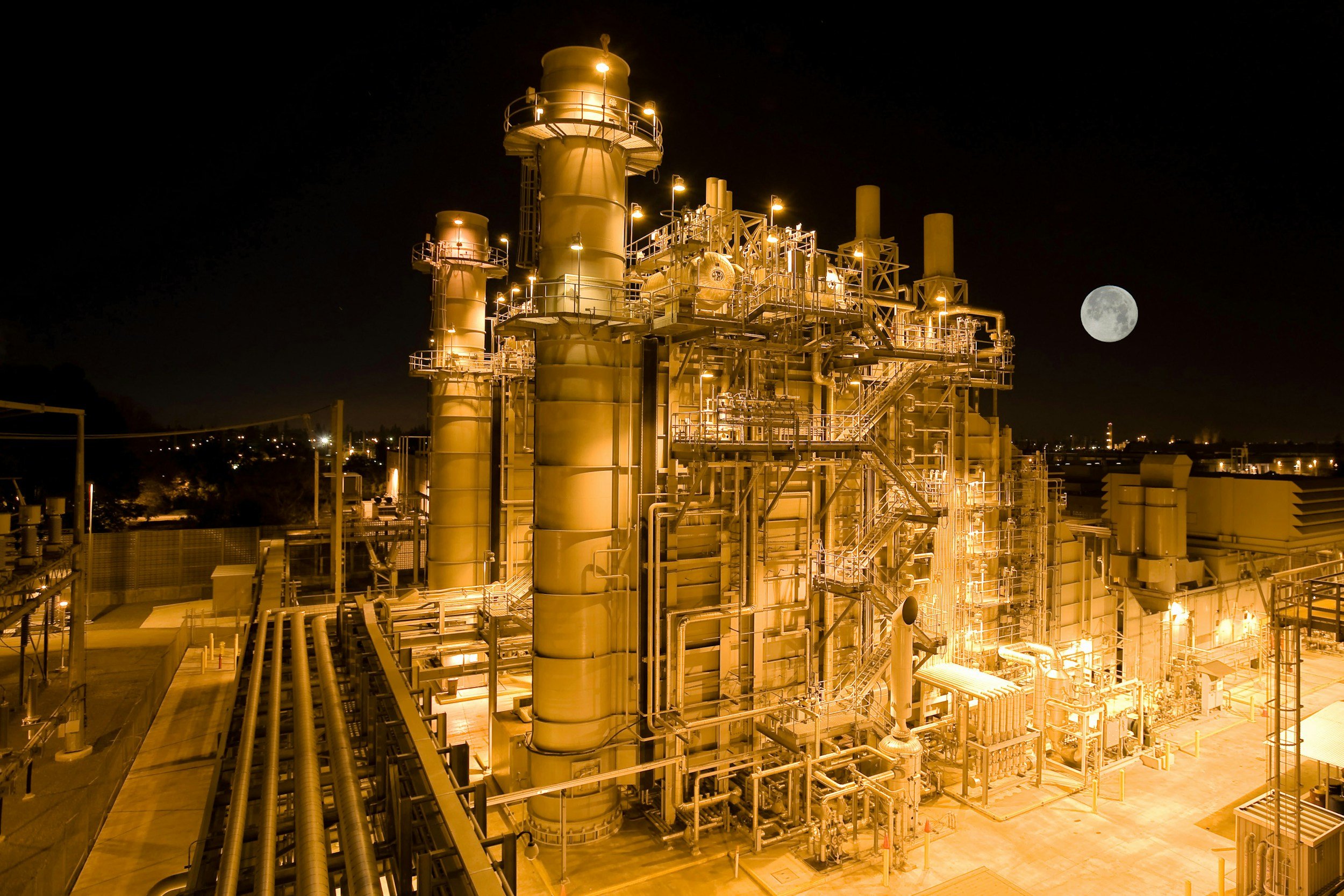 Night view of an industrial facility with large yellow pipes and structures illuminated by bright yellow lights, and a full moon in the dark sky.