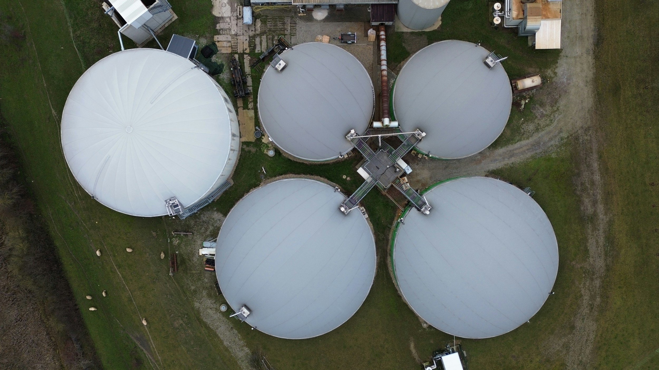 An aerial view of a facility with several large, white, dome-shaped storage tanks connected by pipes and walkways. Surrounding the tanks is grass and gravel pathways.