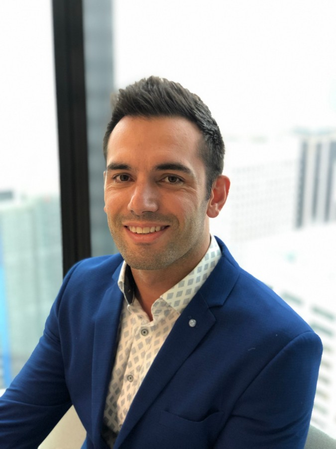 A man in a blue suit jacket smiling in front of a window with a city skyline in the background.