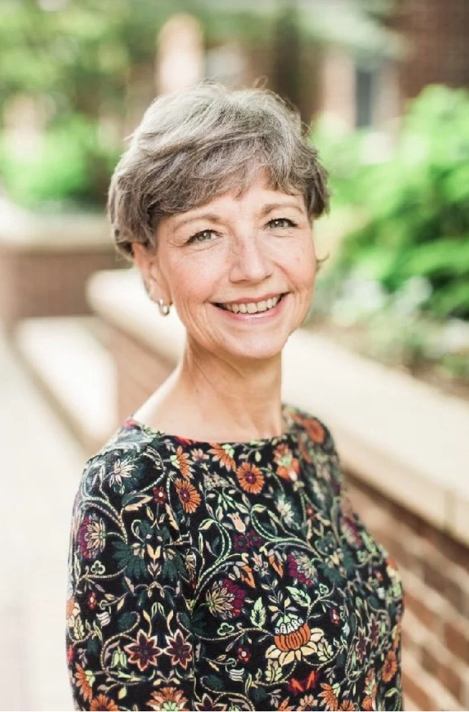 A smiling older woman with short gray hair wearing a black floral embroidered top, standing outdoors with green foliage and a brick wall in the background.