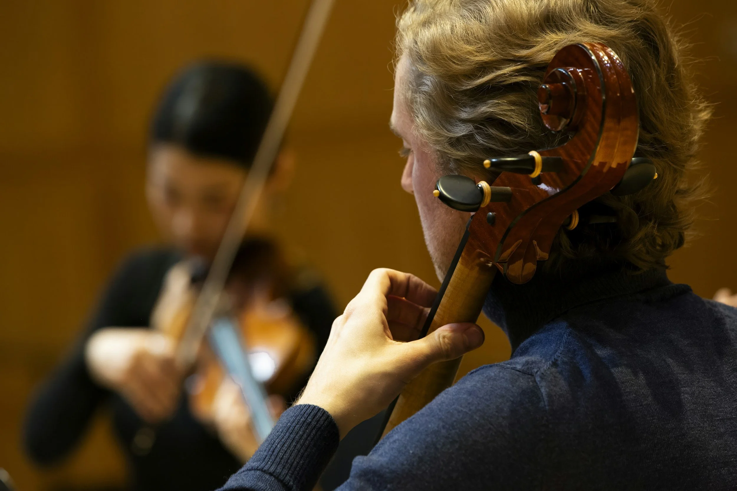 Close-up of a musician with long curly blond hair playing a violin, wearing a headset microphone, in a concert setting.