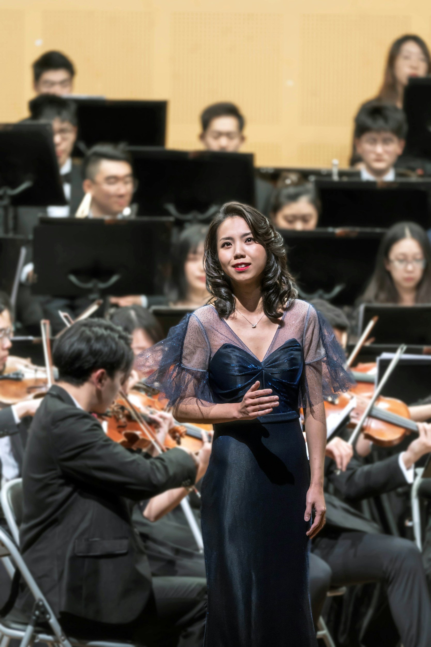 A female singer in a dark blue, sheer evening gown with a bow detail in the front, singing on stage with an orchestra in the background.