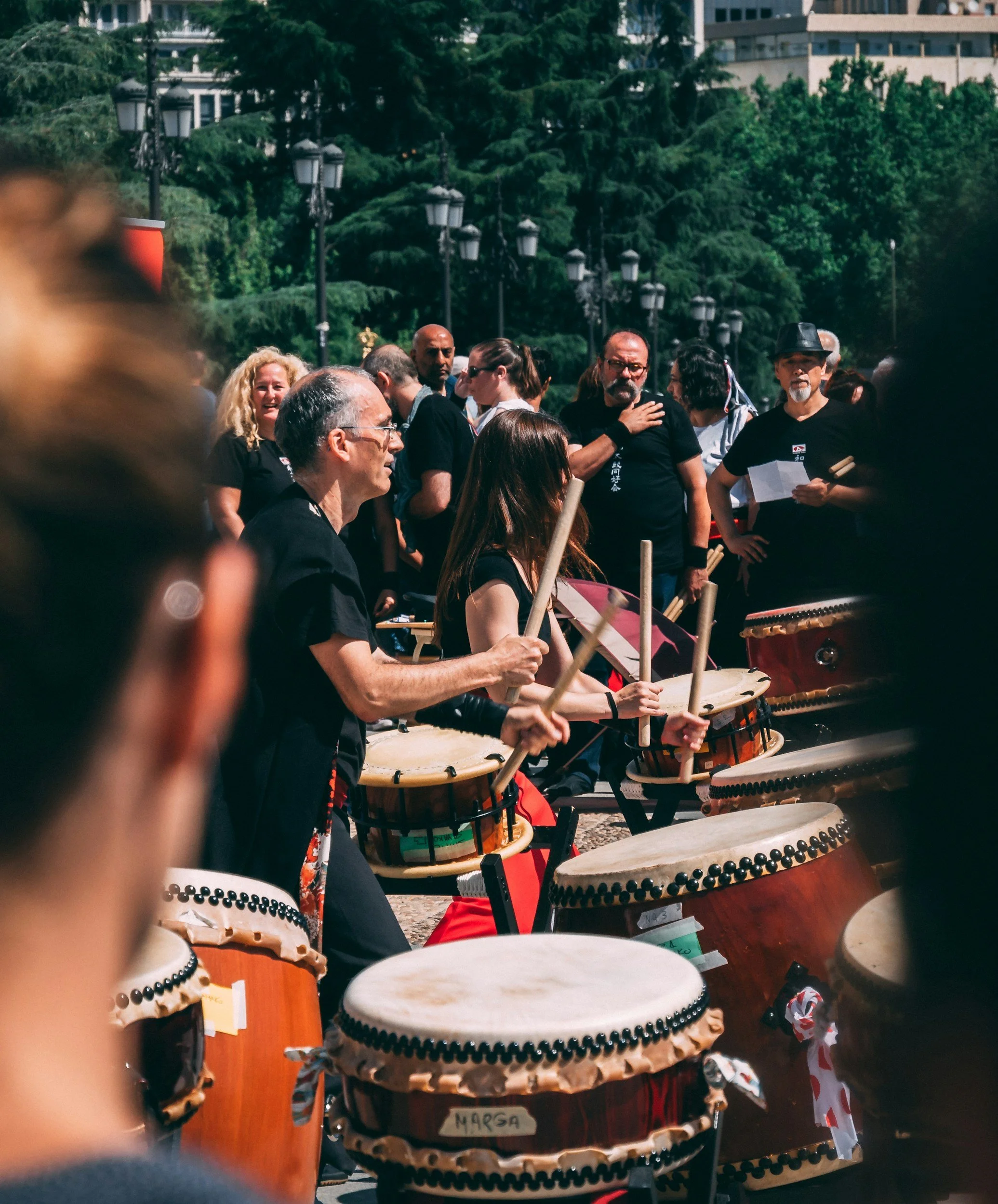 A group of people gather outdoors playing traditional drums, with a backdrop of trees, street lamps, and modern buildings.