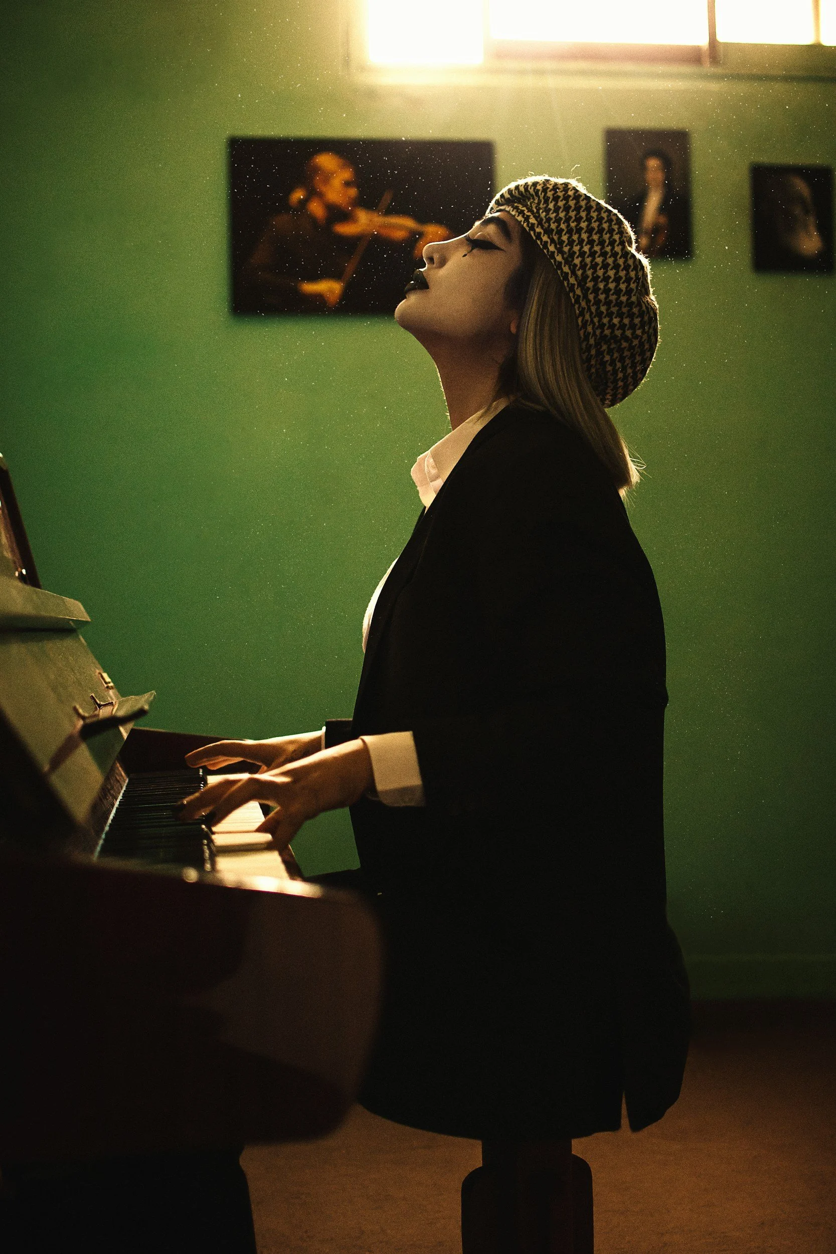 A woman with a houndstooth beret and dark makeup playing a grand piano in a room with green walls and framed photographs, backlit by sunlight from a window.