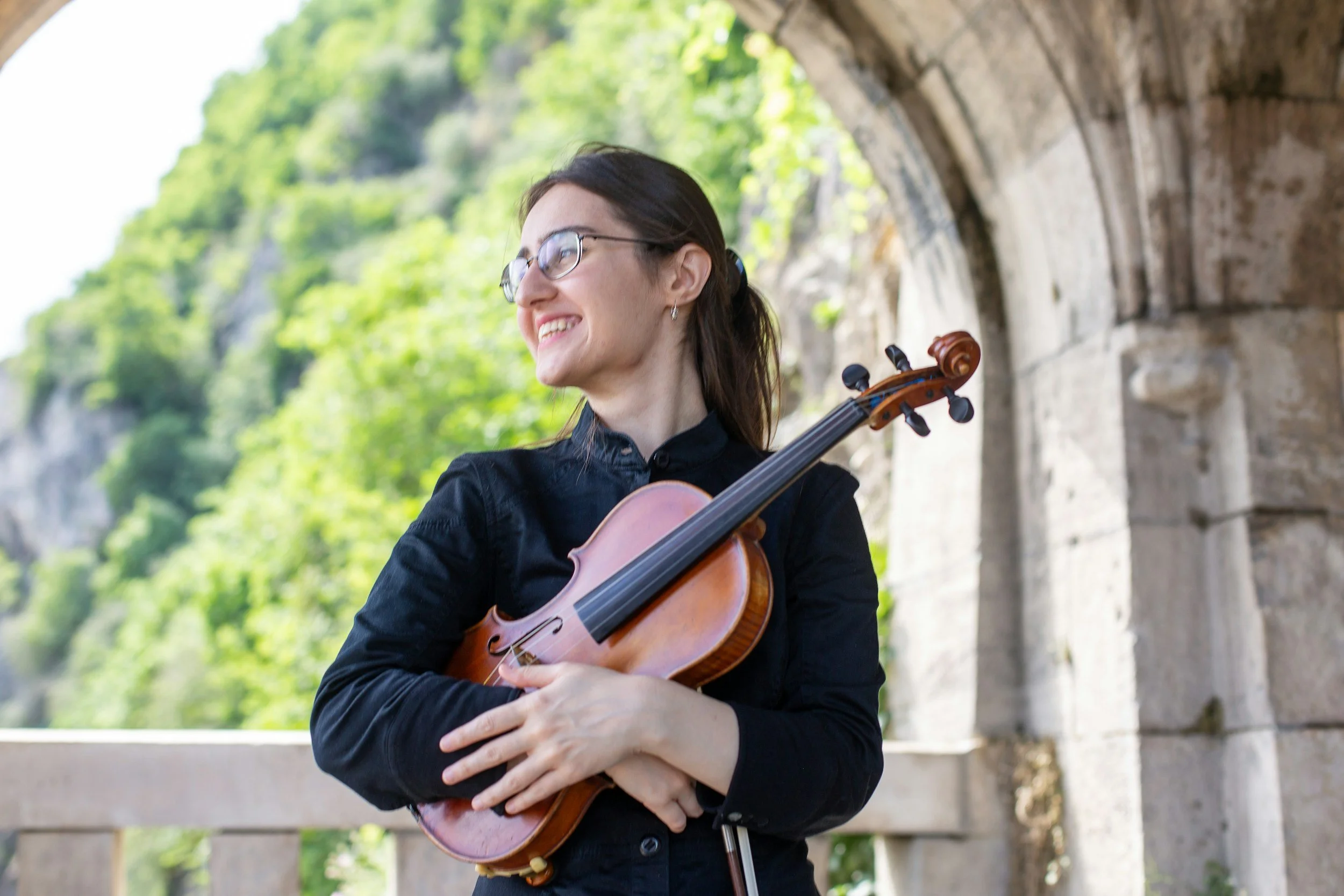 A woman wearing glasses and a black shirt, smiling while holding a violin, outdoors with lush green trees and stone archway in the background.