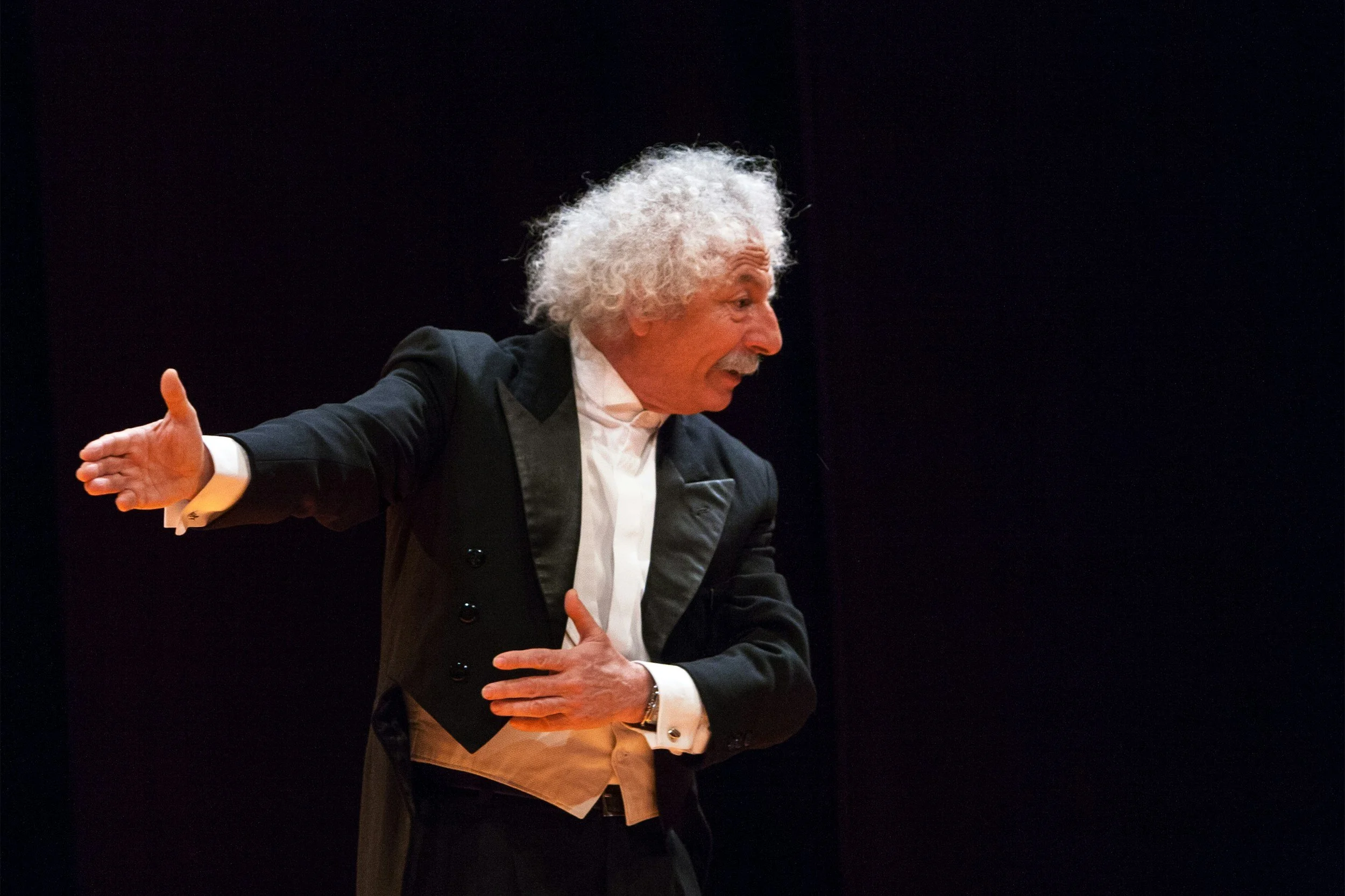 A man with curly gray hair dressed in a formal black tuxedo with white shirt and black bow tie, speaking or performing on stage against a dark background.