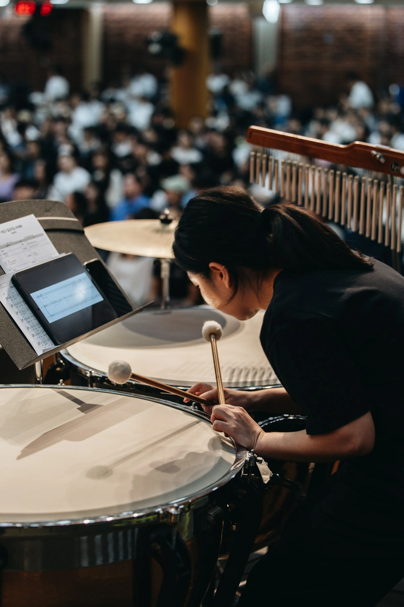 A musician playing a steelpan drum at a concert or event with a large seated audience in the background.