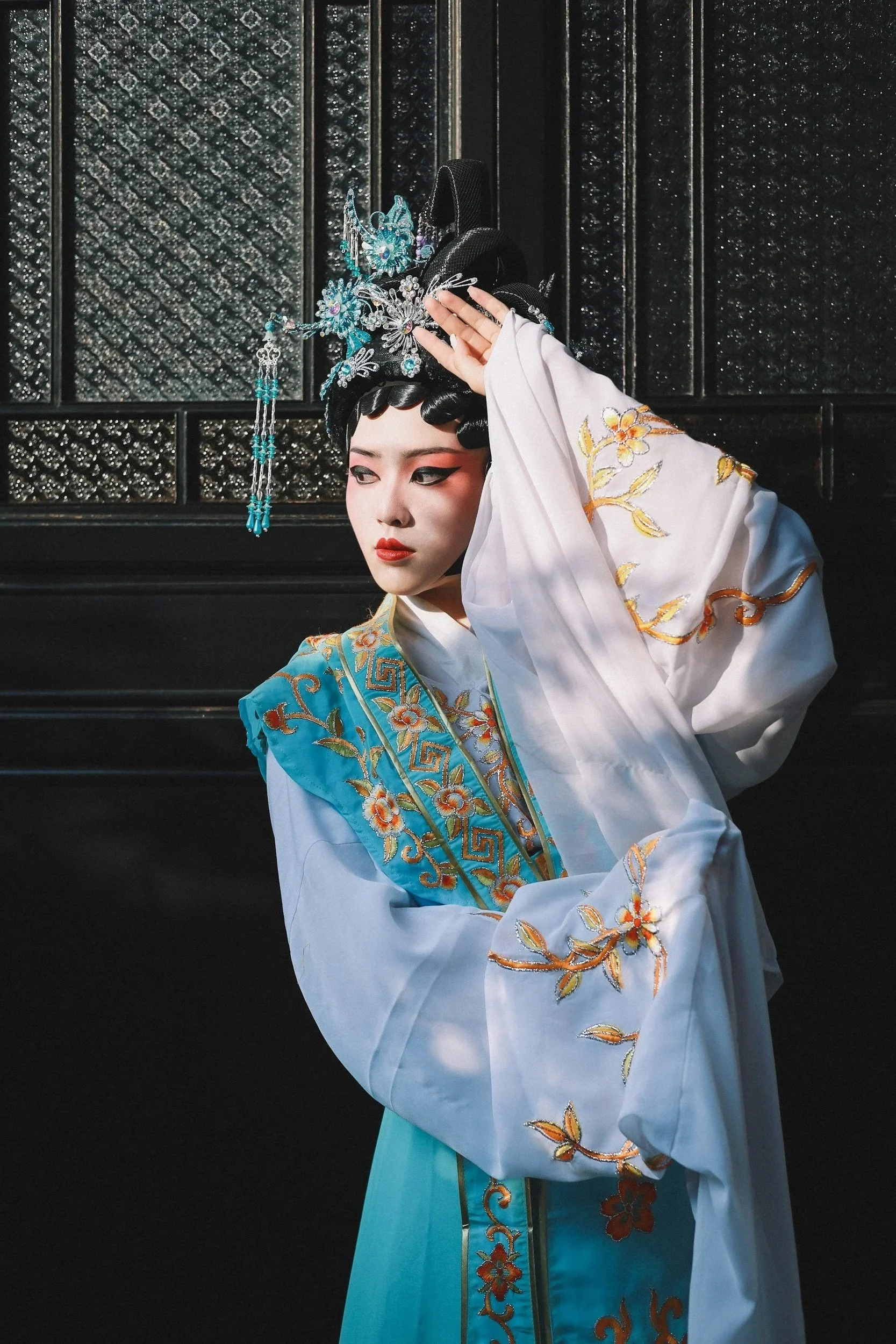 A woman dressed in traditional Chinese opera costume with intricate embroidery, wearing elaborate headpieces adorned with jewelry and blue gemstones, standing against a dark textured background.