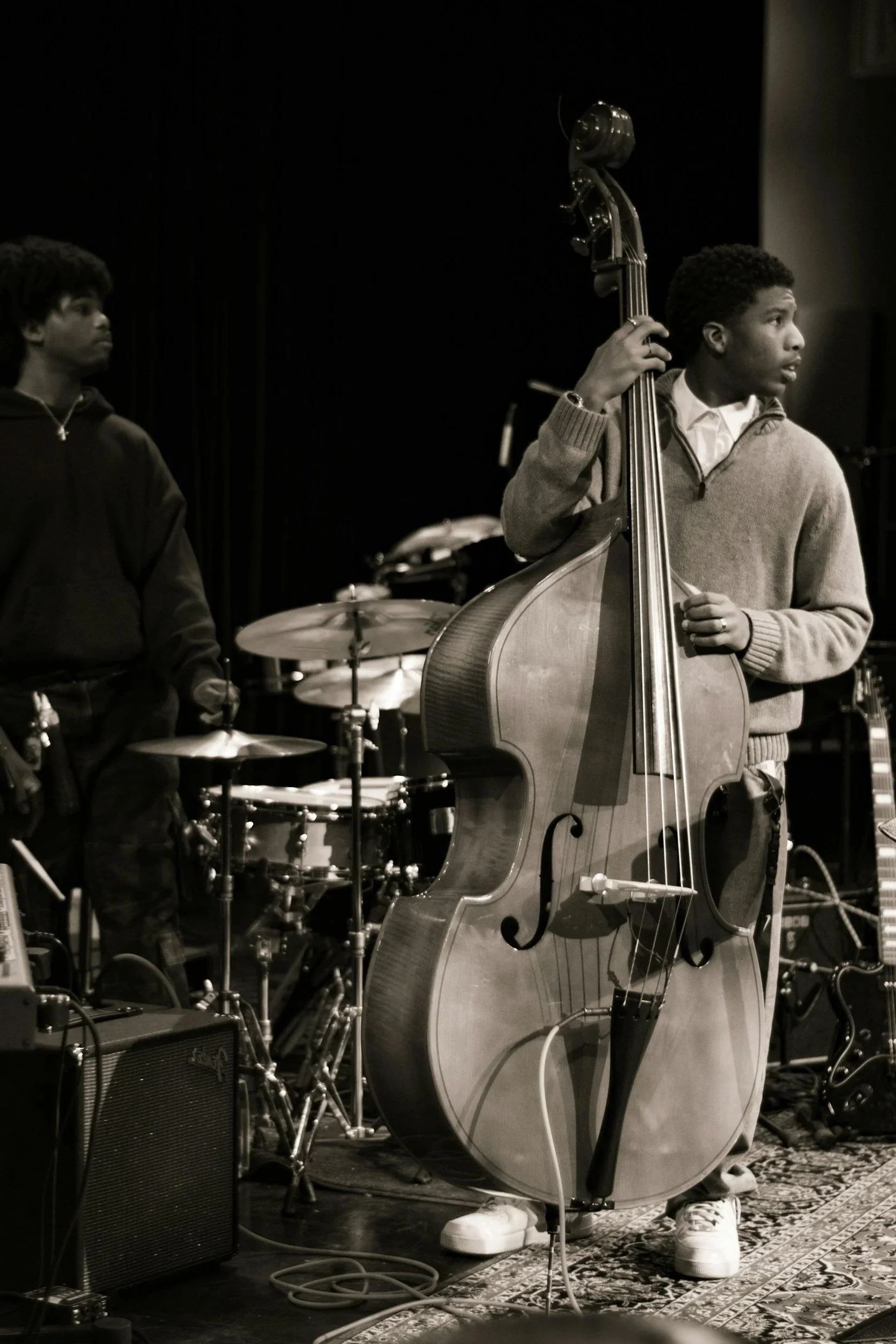 Two musicians on stage, one playing drums and the other playing an upright bass, with musical equipment and a dark background.