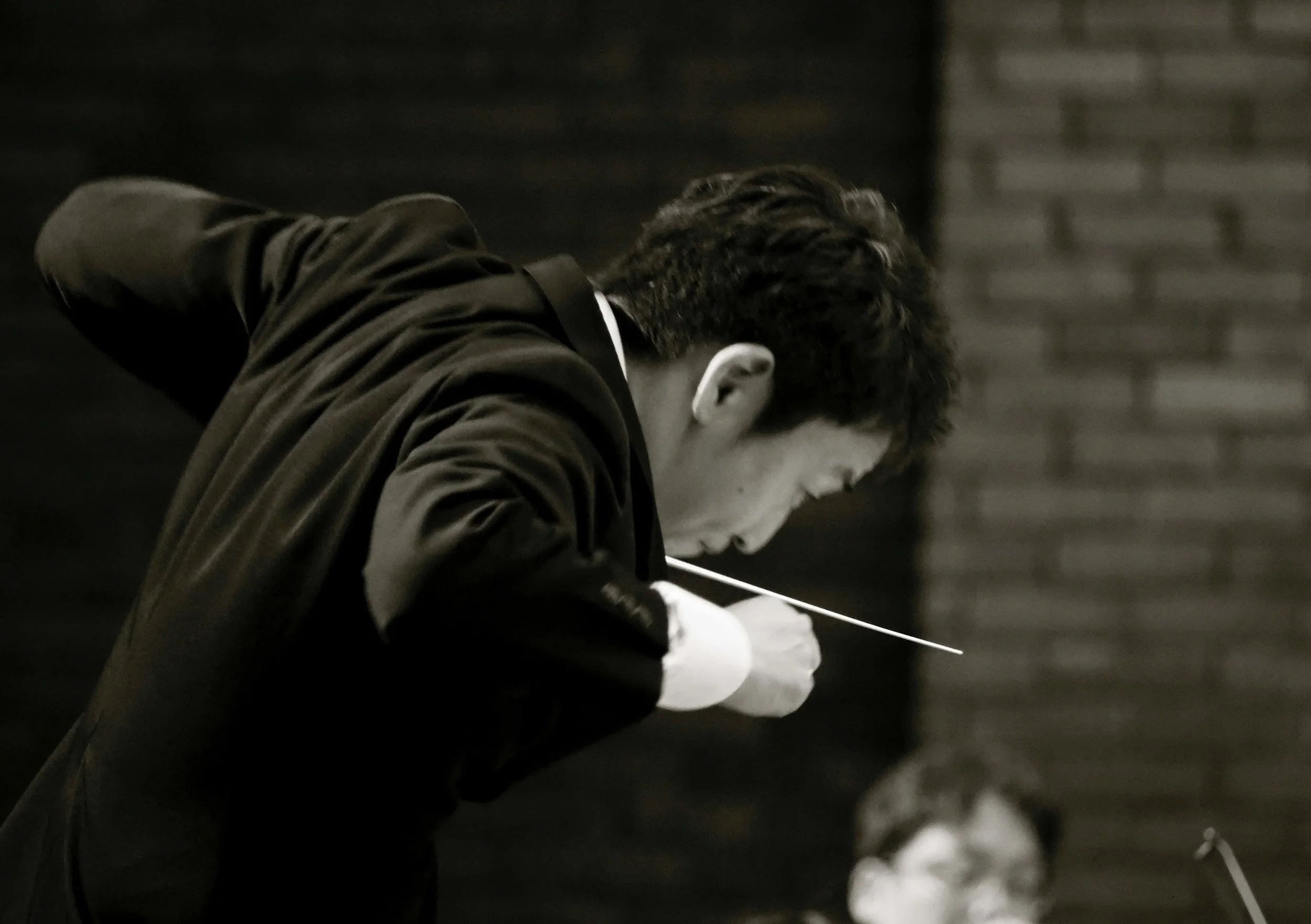 A young man with dark hair, wearing a dark jacket, is leaning over a table, writing or drawing on paper with a pencil.