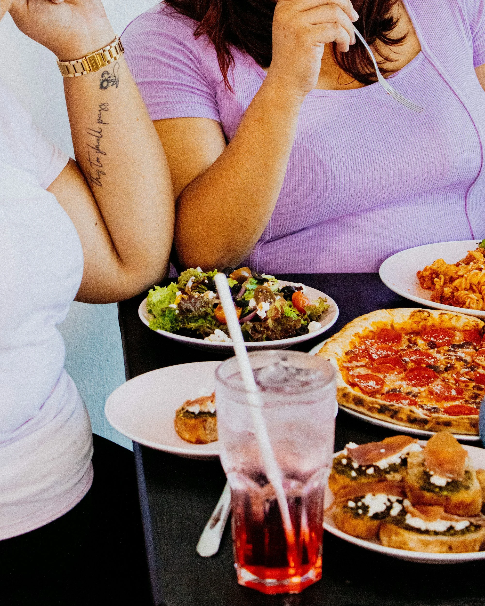 A table with various food items including pizza, pasta, salads, and drinks. A person is holding a fork, and there are plates of food and a glass of cola with a straw in the foreground.