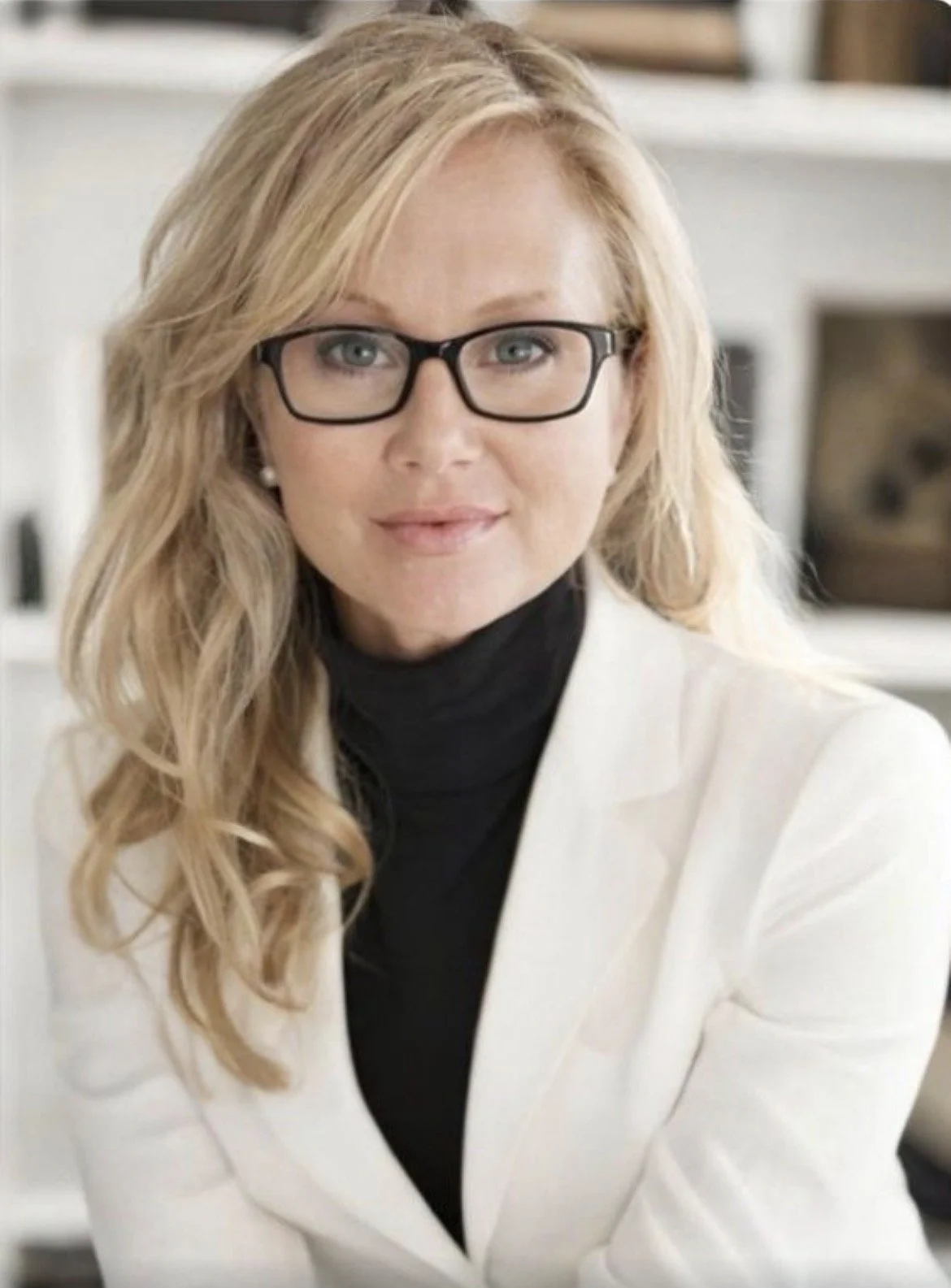 A woman with blonde wavy hair, glasses, wearing a white blazer over a black turtleneck, sitting indoors with a blurred bookshelf in the background.