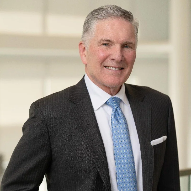 A smiling middle-aged man in a dark suit, white shirt, and blue tie with a patterned background.