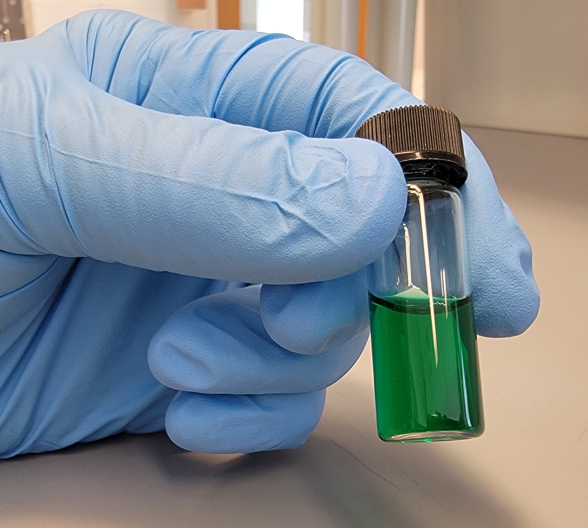 A person wearing blue medical gloves holds a glass container with a black lid. The container is filled with a clear green liquid. The person is in a setting that looks like a laboratory or medical environment.
