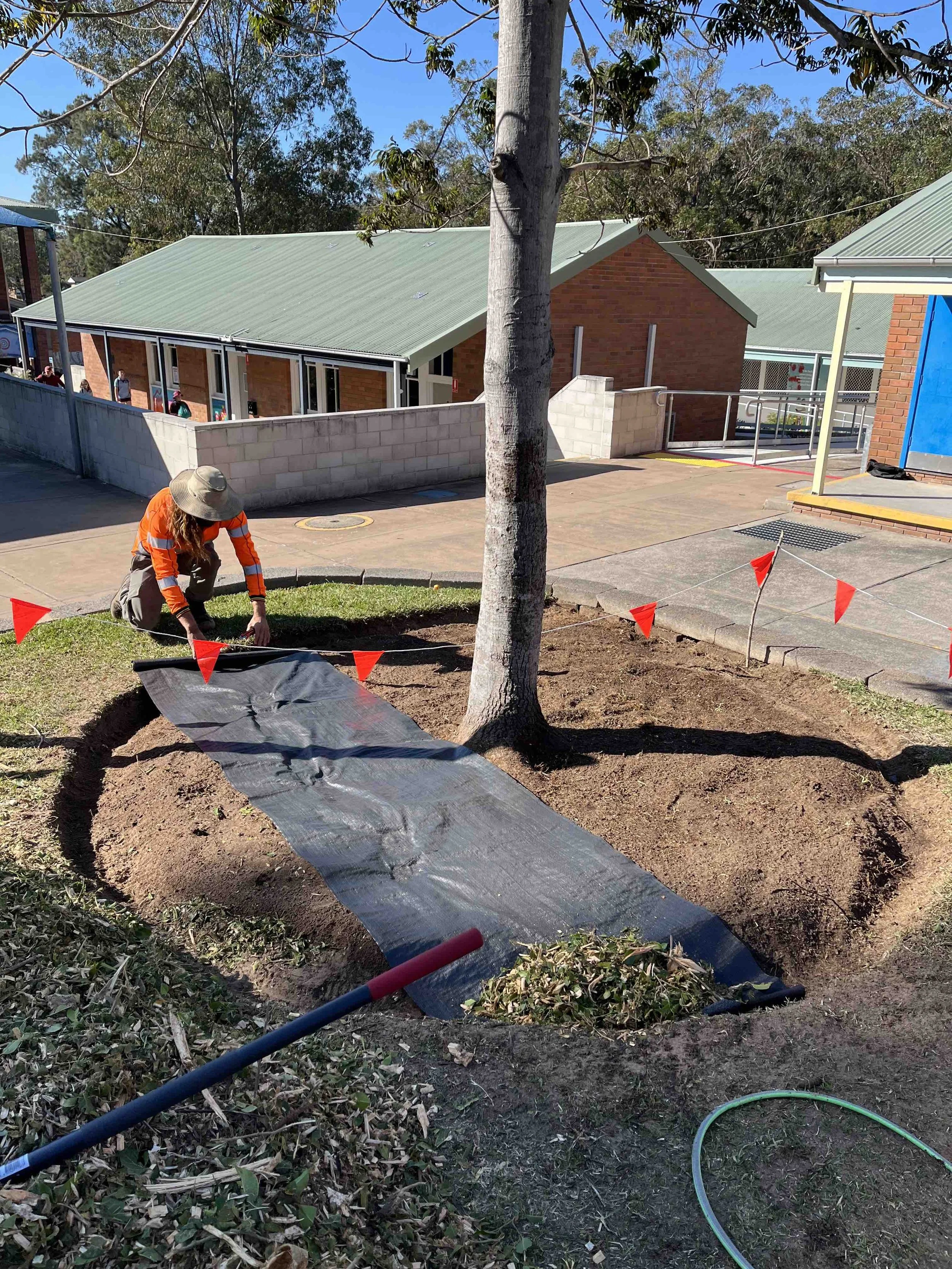 Person planting a tree in a garden with a black cover underneath, surrounded by red flags, in front of a building with a green roof.