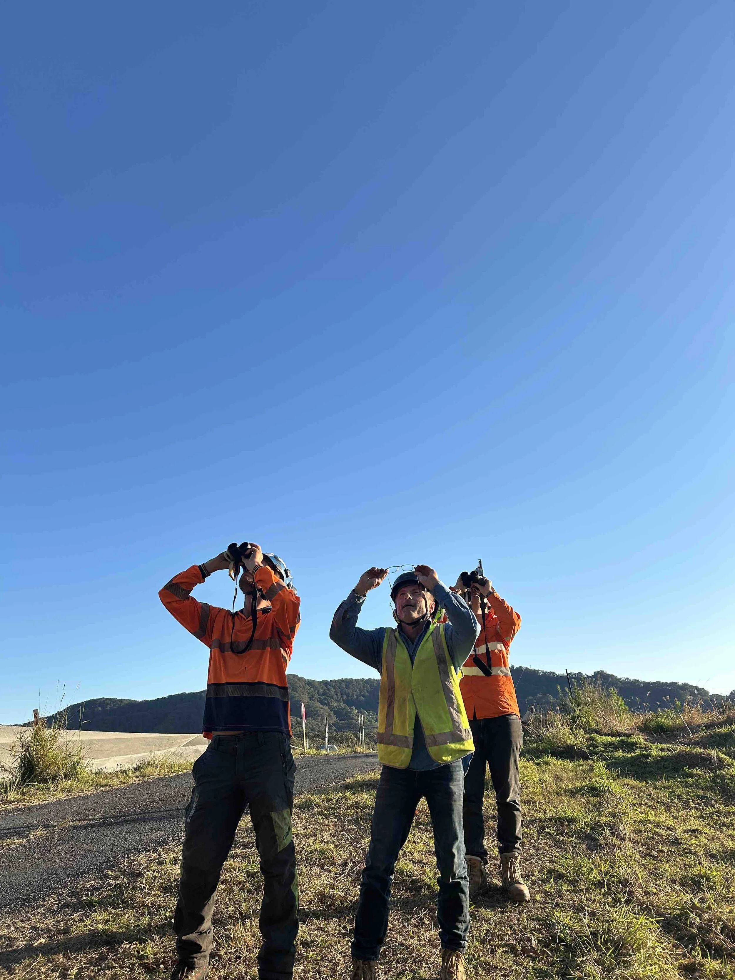 Four people standing outdoors on a clear day, some wearing high-visibility vests and holding binoculars, with hilly terrain and blue sky in the background.