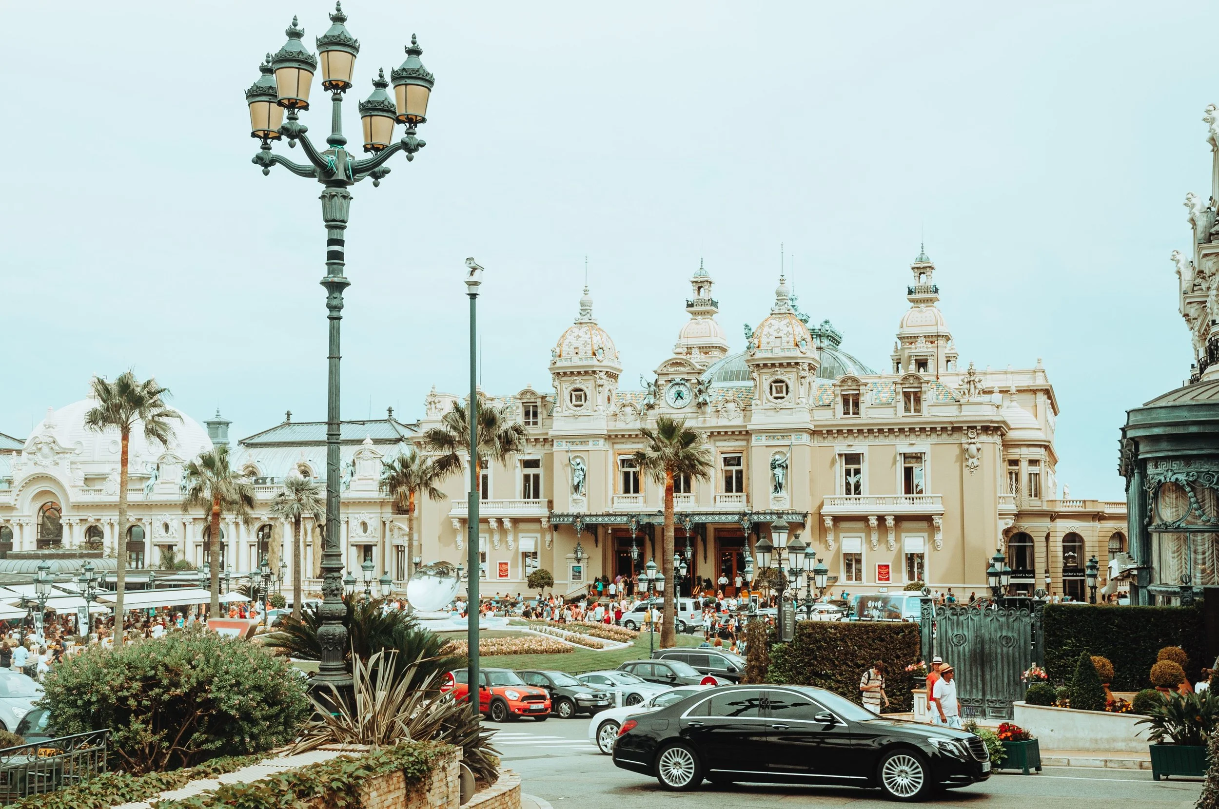 A large ornate building with a clock tower, surrounded by palm trees, and an outdoor crowd gathered in front. In the foreground, there is a parking lot with several cars, including a black sports car, and street lamps. The sky is overcast.