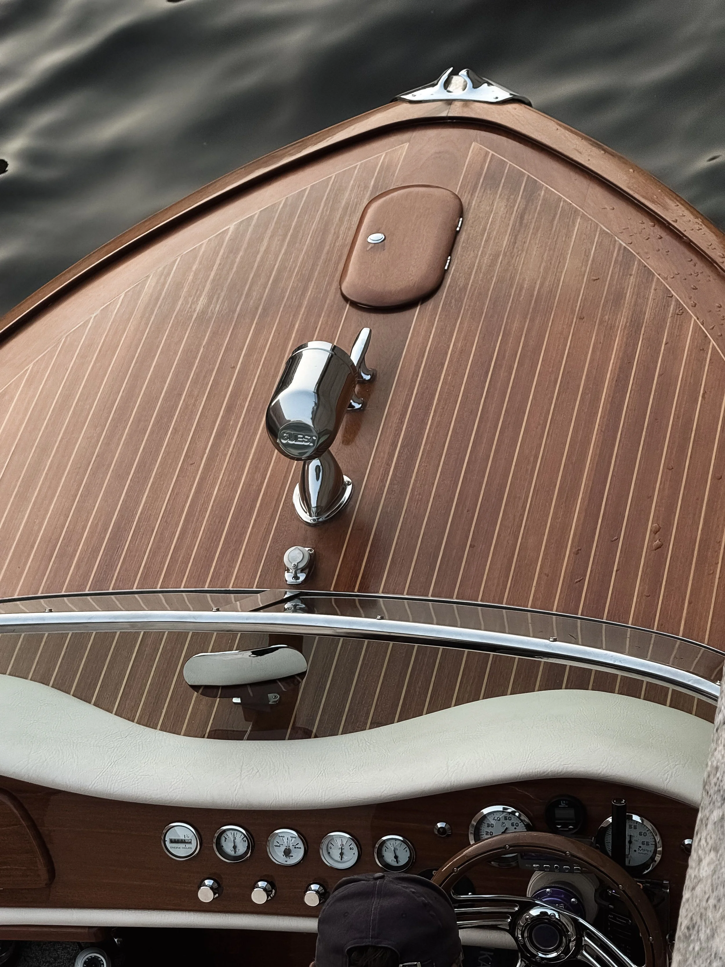 The image shows the dashboard of a boat with analog gauges, a steering wheel, and a navigation gear. The boat's deck appears to be wooden with a brown finish, and the water is visible in the background.