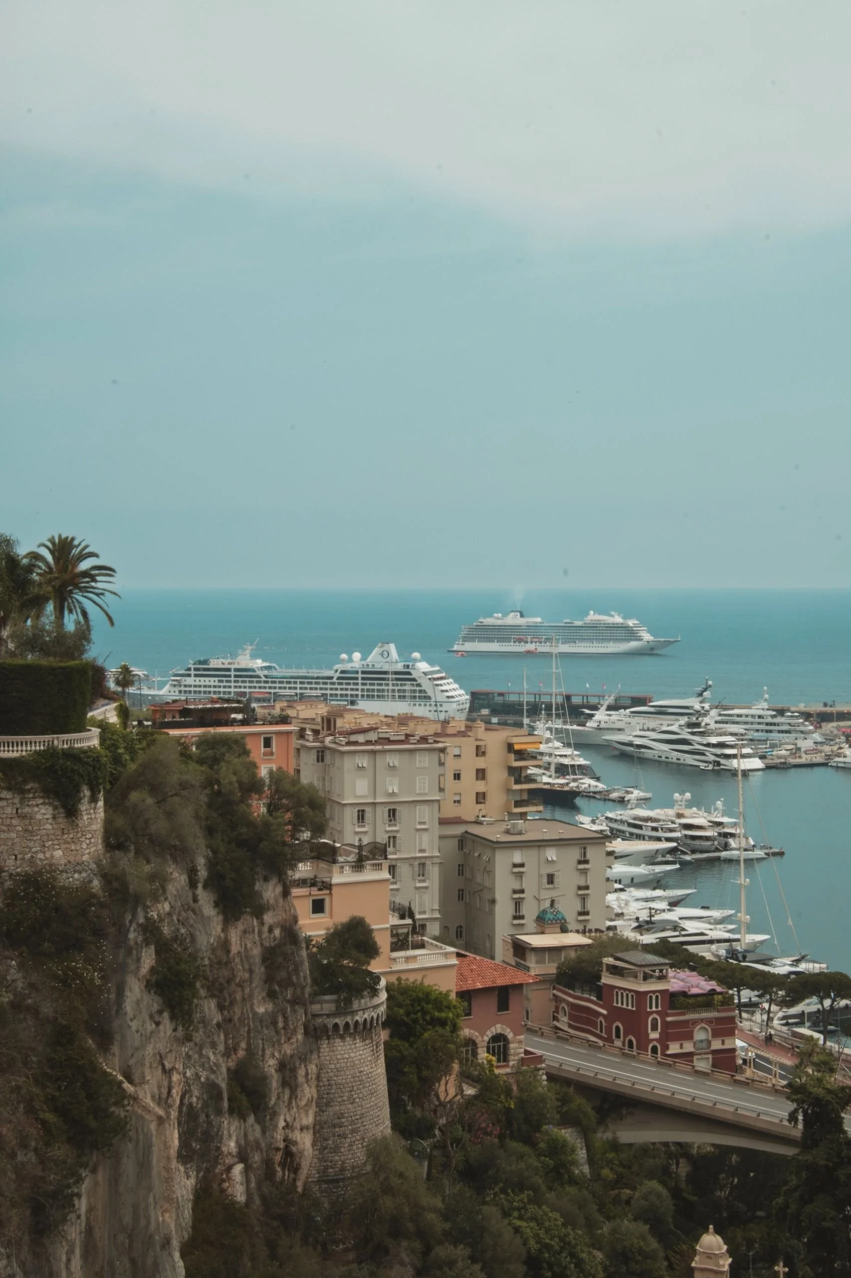 A hillside with historic architecture and numerous yachts anchored in a harbor, with a large cruise ship visible in the background at sea.