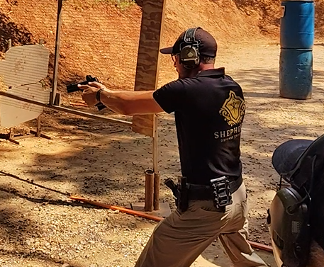 A man wearing a black shirt with sheriff insignia, tan pants, and ear protection is aiming a handgun at a shooting range. He is in a shooting stance with a blue barrel and brick wall in the background.