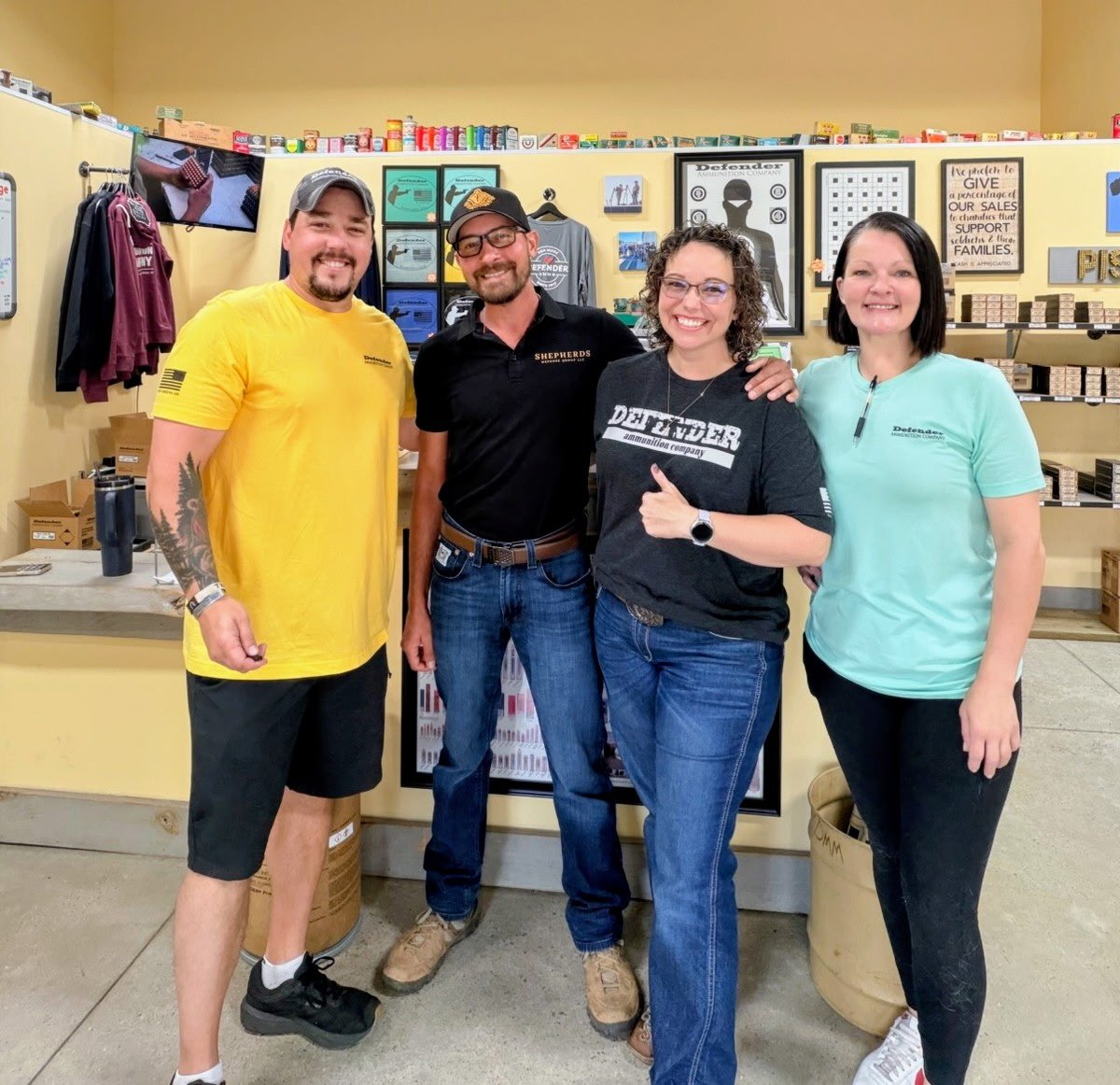 Four people standing inside a store, smiling at the camera. Two women and two men, wearing casual clothing, with shelves and signs in the background.