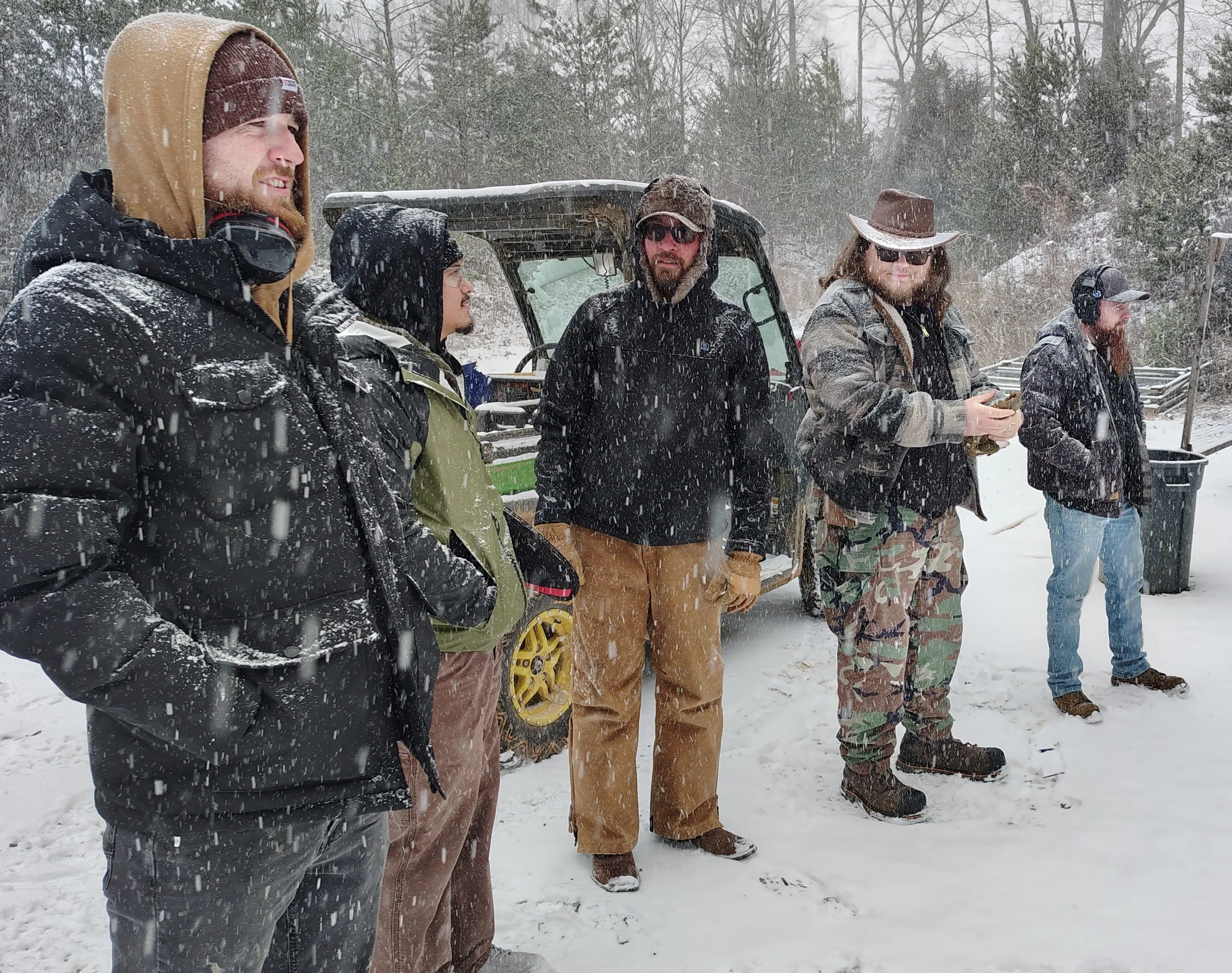 Group of five people standing in a snowy outdoor area with trees and a wooden fence in the background. They are dressed in winter clothing, including jackets, hats, gloves, and boots, and are facing to the right. Heavy snowfall is visible in the scene.