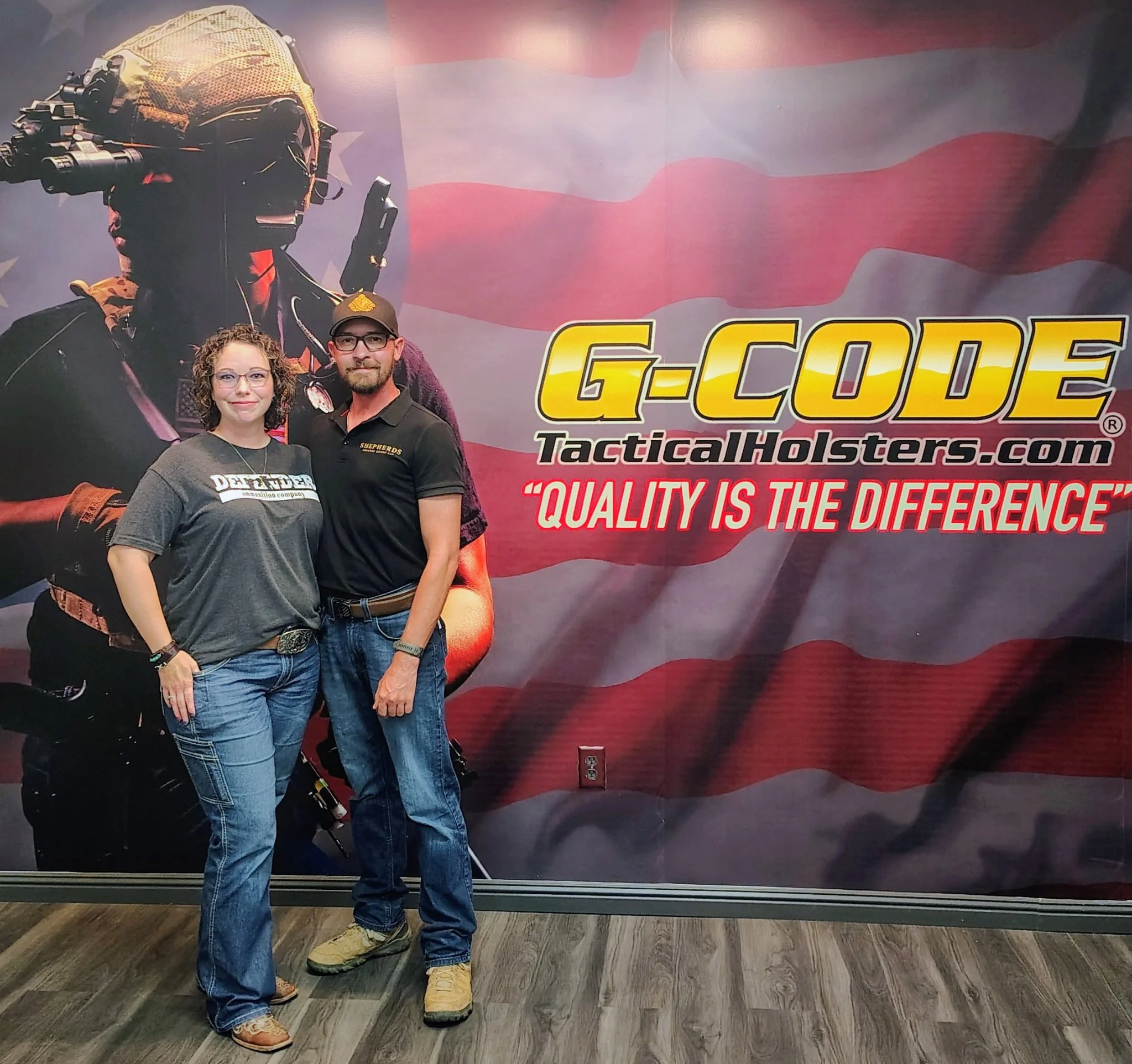Two people standing in front of a large American flag mural with the logo and website for G-Code Tactical Holsters. One woman with glasses and curly hair, and a man with a beard wearing glasses, a cap, and a black polo shirt. Both are smiling and posing for the photo.