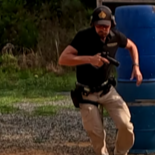 A man running outdoors near a blue structure, wearing a black cap, black shirt, and beige pants.