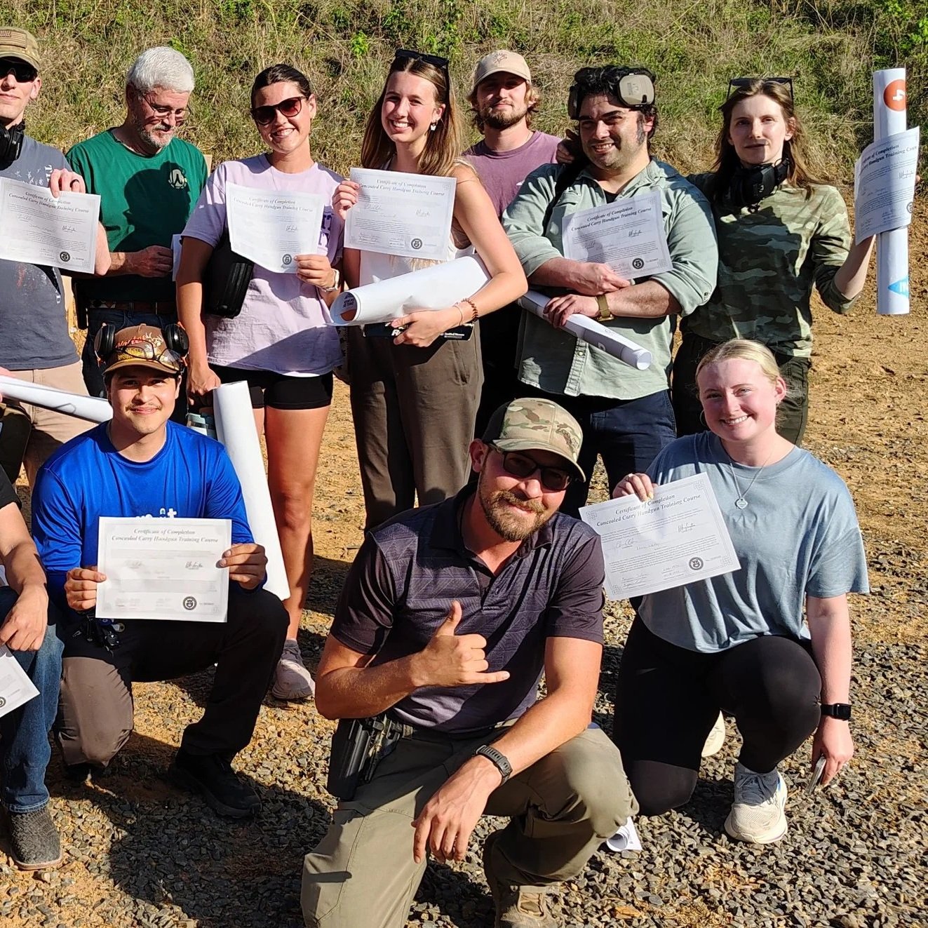 Group of people outdoors holding certificates, smiling, and posing for a photo on rocky terrain with grass behind them.