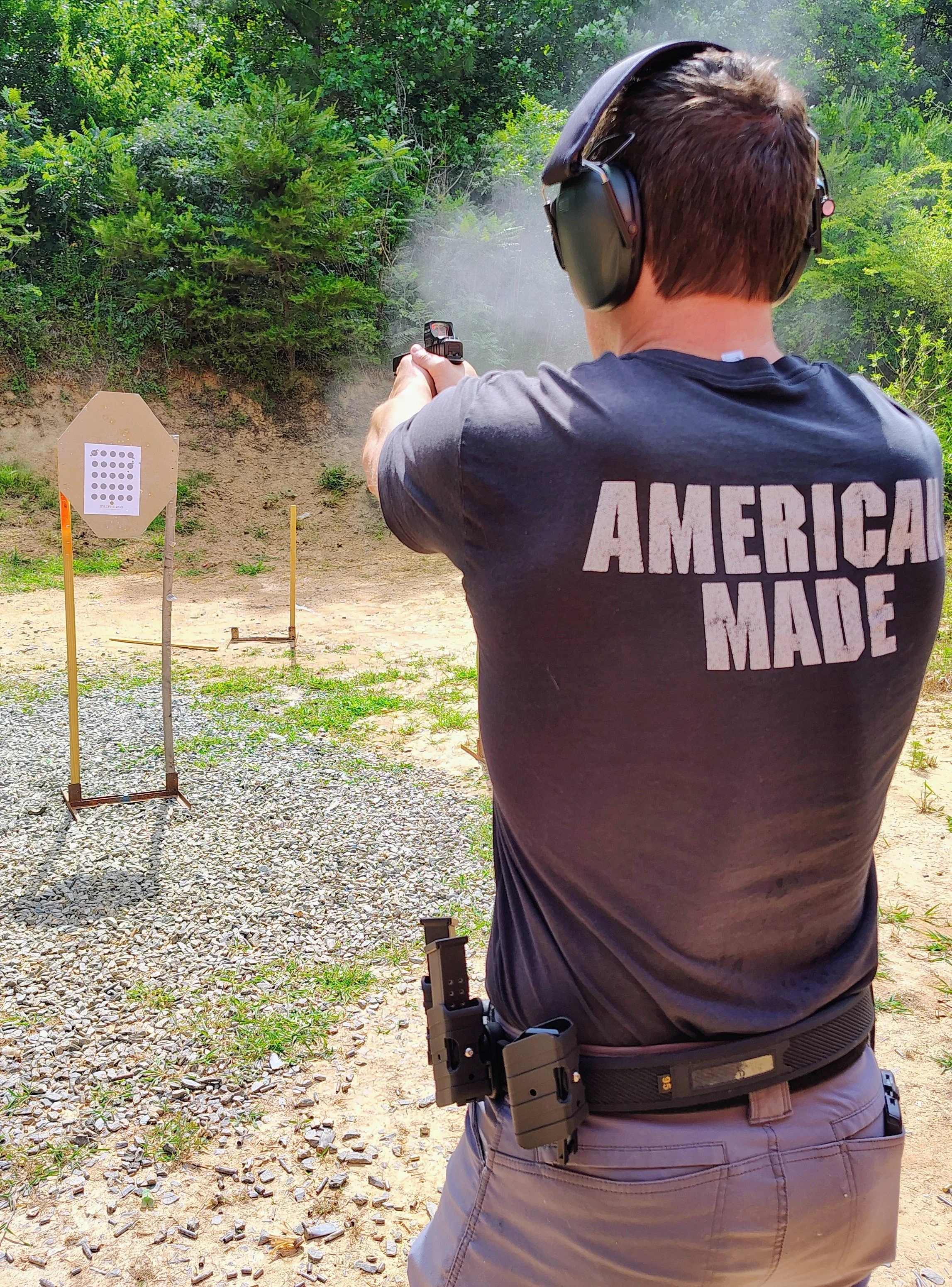 A person with short brown hair wearing hearing protection and a dark T-shirt with "AMERICA MADE" written on the back is shooting a handgun at an outdoor shooting range. The person has a handgun holstered on their waist and is aiming at a paper target with a grid of black dots, set up in front of green foliage.