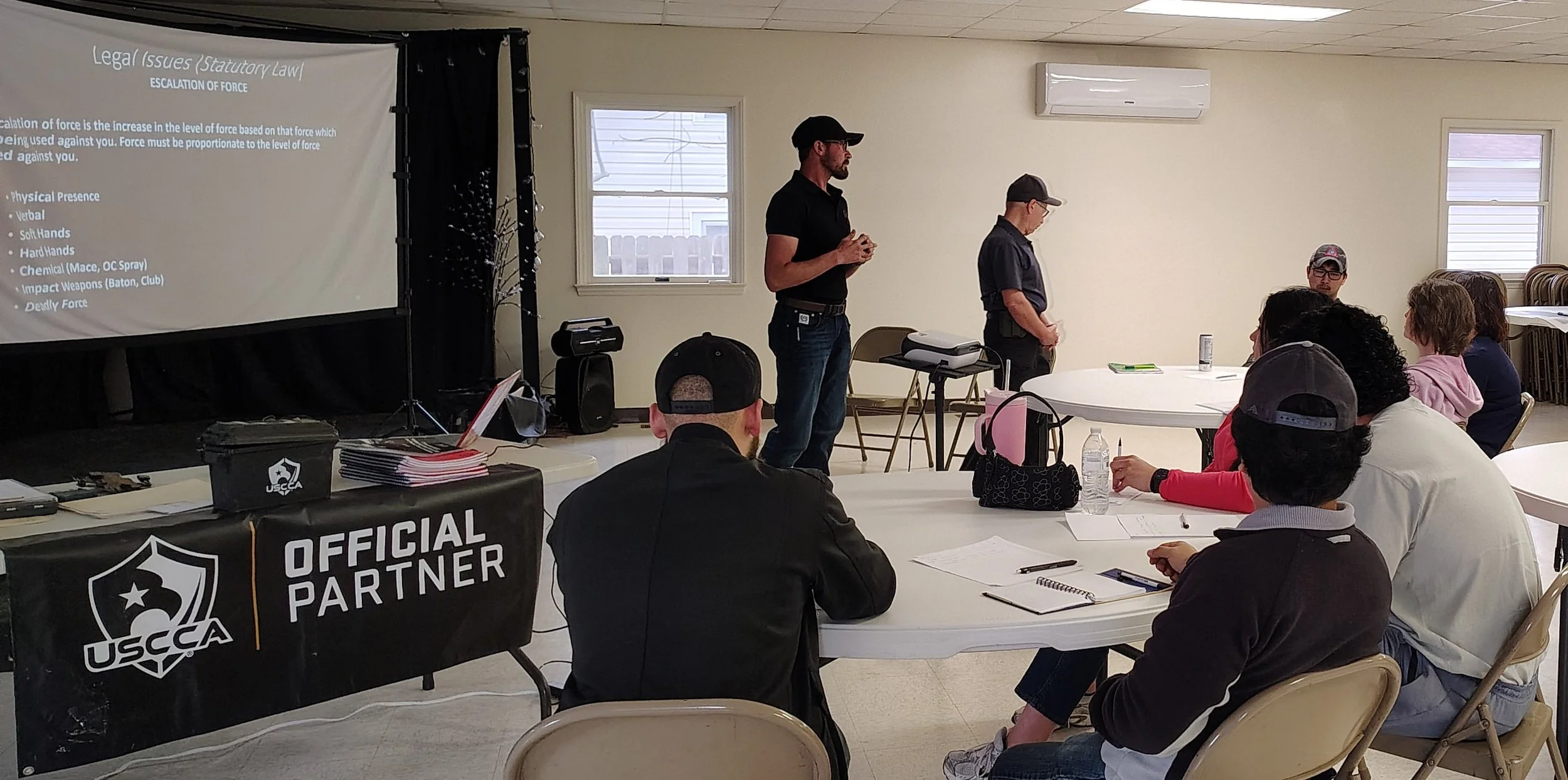 A person presenting in front of a group during a training session about legal issues and escalation of force, with participants seated at round tables listening and taking notes.