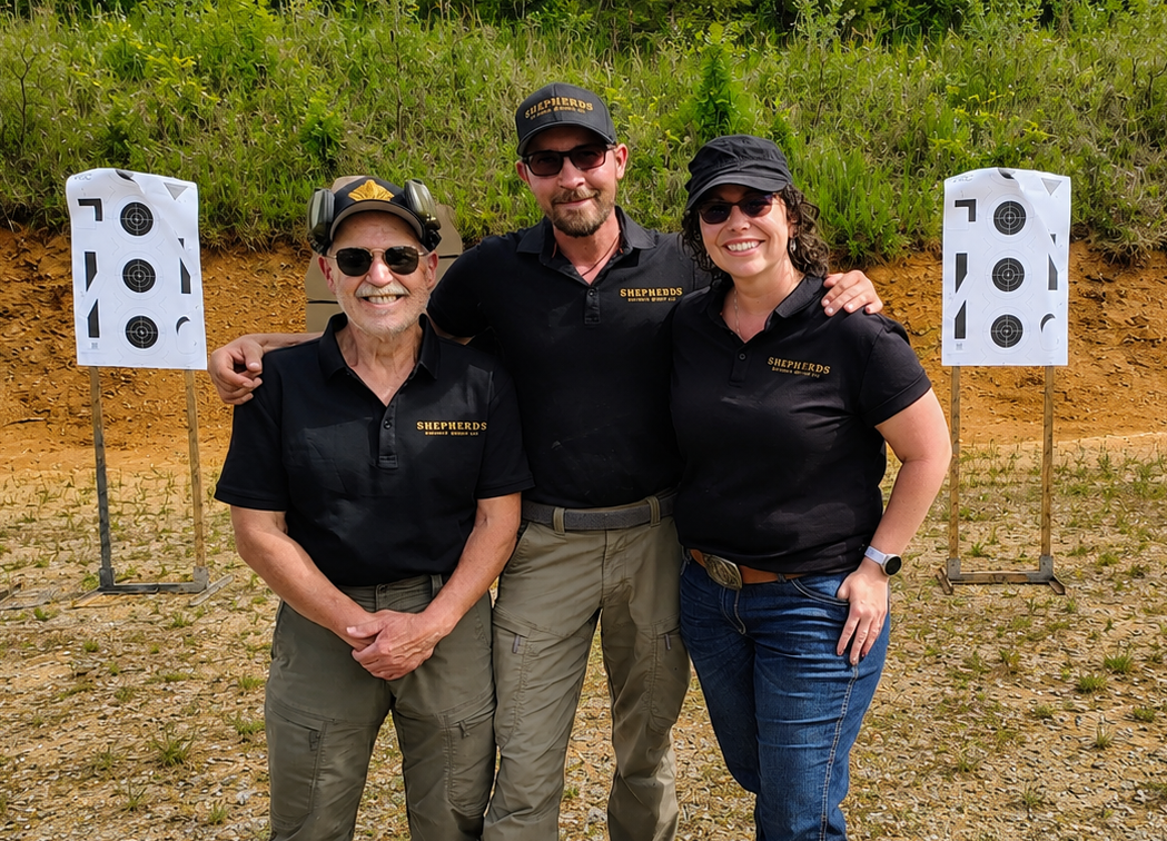 Three people standing outdoors in front of shooting targets, wearing matching black shirts with 'Shepherds' logo, smiling and posing for the photo.