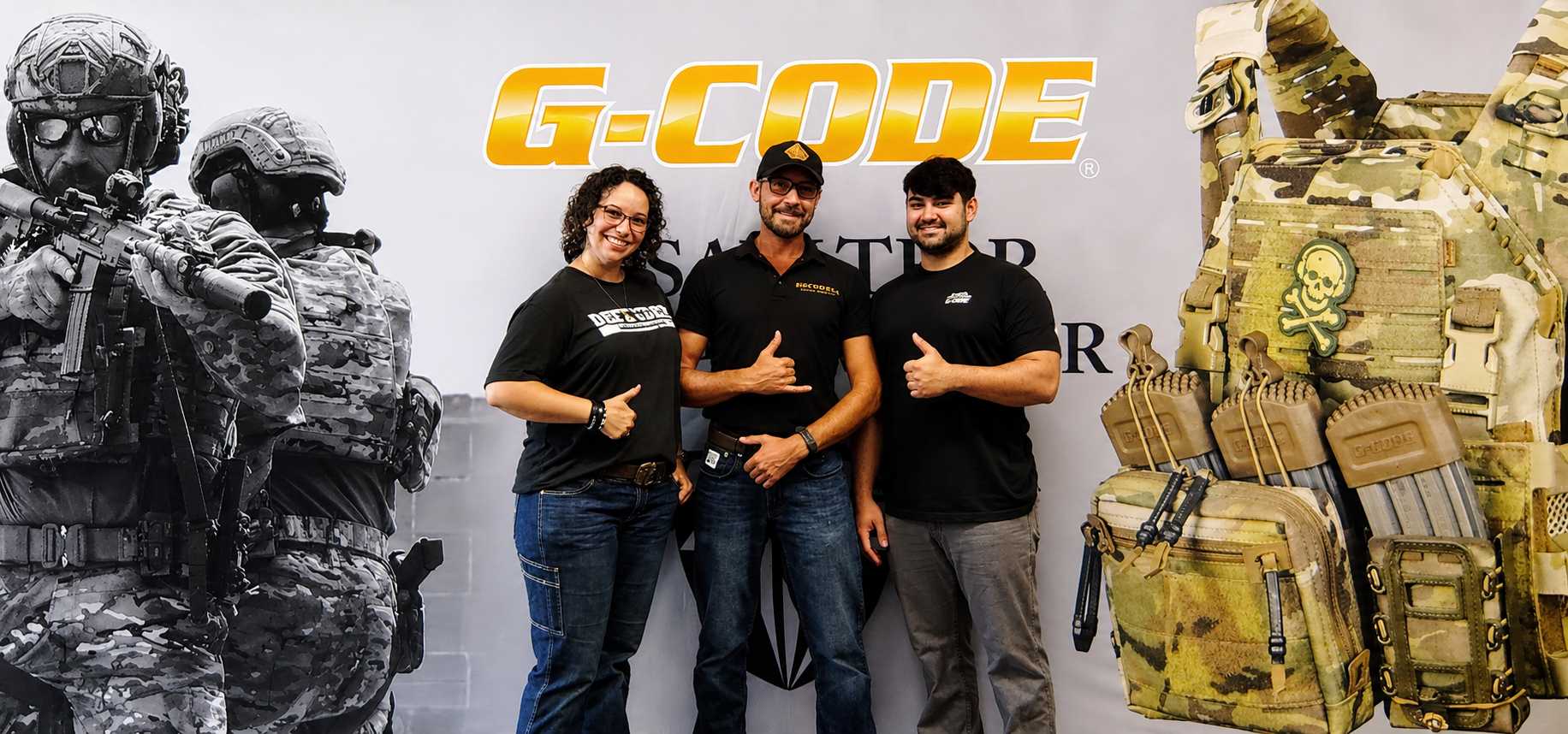 Three people smiling and posing in front of a G-CODE banner with tactical gear and military-style clothing on display.