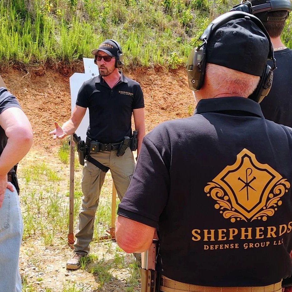 A man in a black shirt with 'SHEPHERDS' on it, is instructing others at an outdoor shooting range. He is wearing protective ear and eye gear, standing in front of a white target, with grassy and dirt terrain in the background.