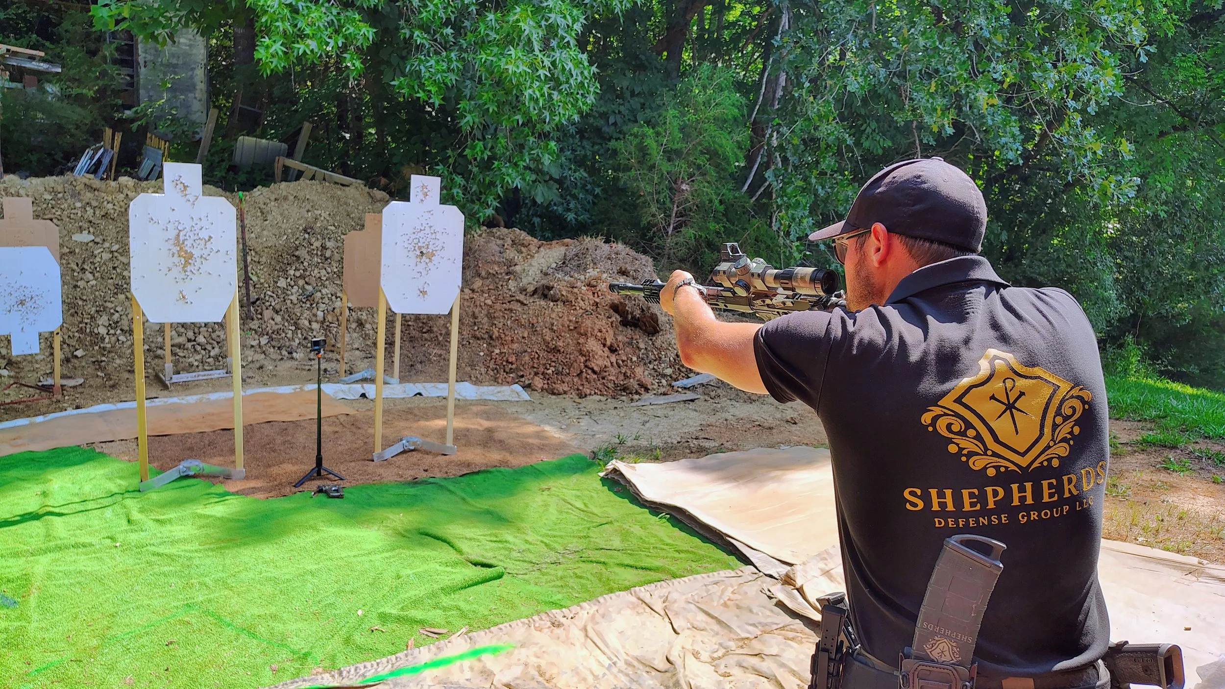 A man wearing a cap and a black shirt with gold lettering and a shield logo is aiming a rifle at outdoor shooting targets. The targets are set on wooden stands against a background of trees and dirt mounds.