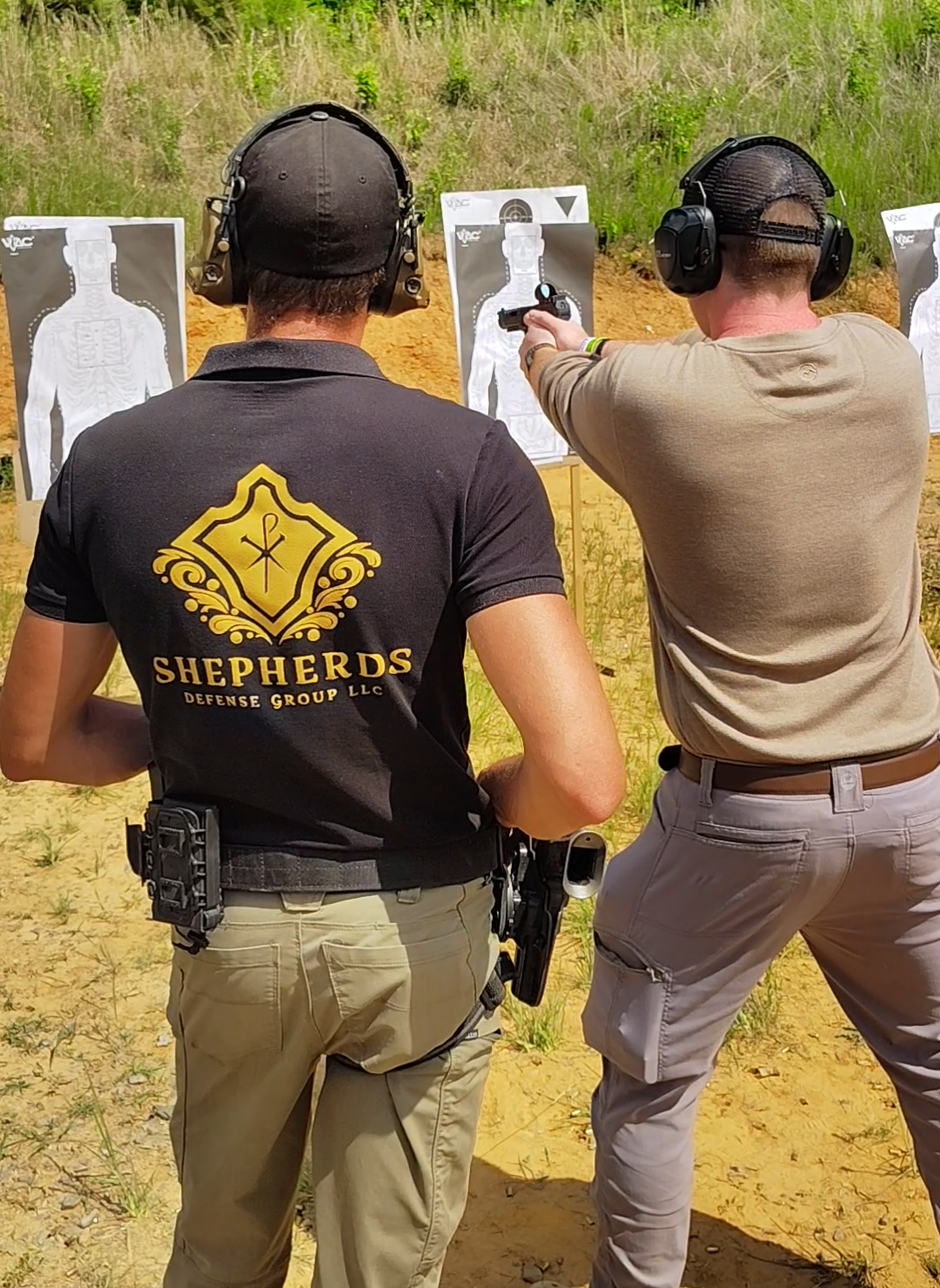 Two men at an outdoor shooting range with paper target targets, one wearing a black shirt with the Sheperds Defense Group logo, and the other in a tan shirt aiming a handgun.