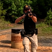 Person in black shirt and tan pants aiming a handgun outdoors
