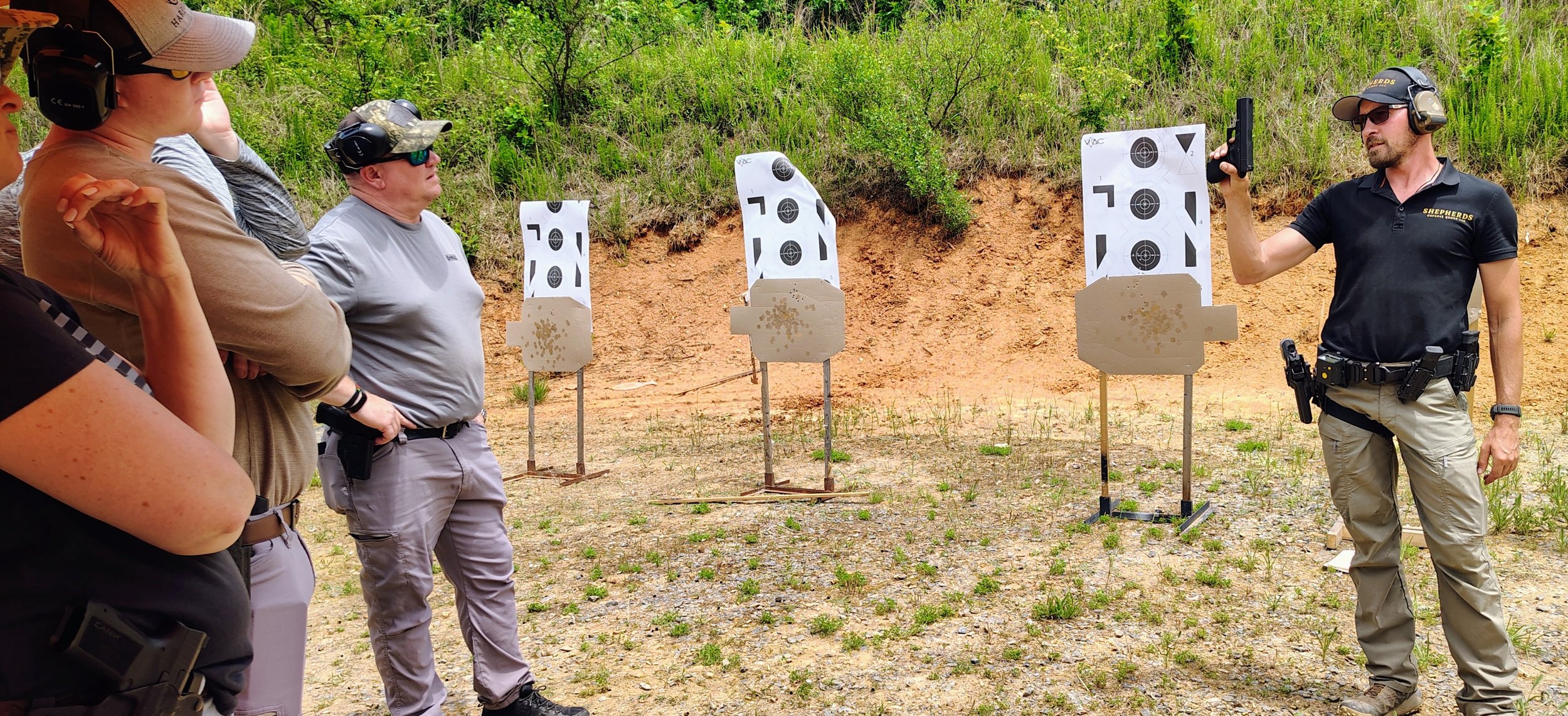 Group of people at an outdoor shooting range, standing near paper target stands with multiple targets, with a man in a black shirt and tan pants explaining something, holding a handgun, and the others listening, surrounded by greenery and dirt ground.