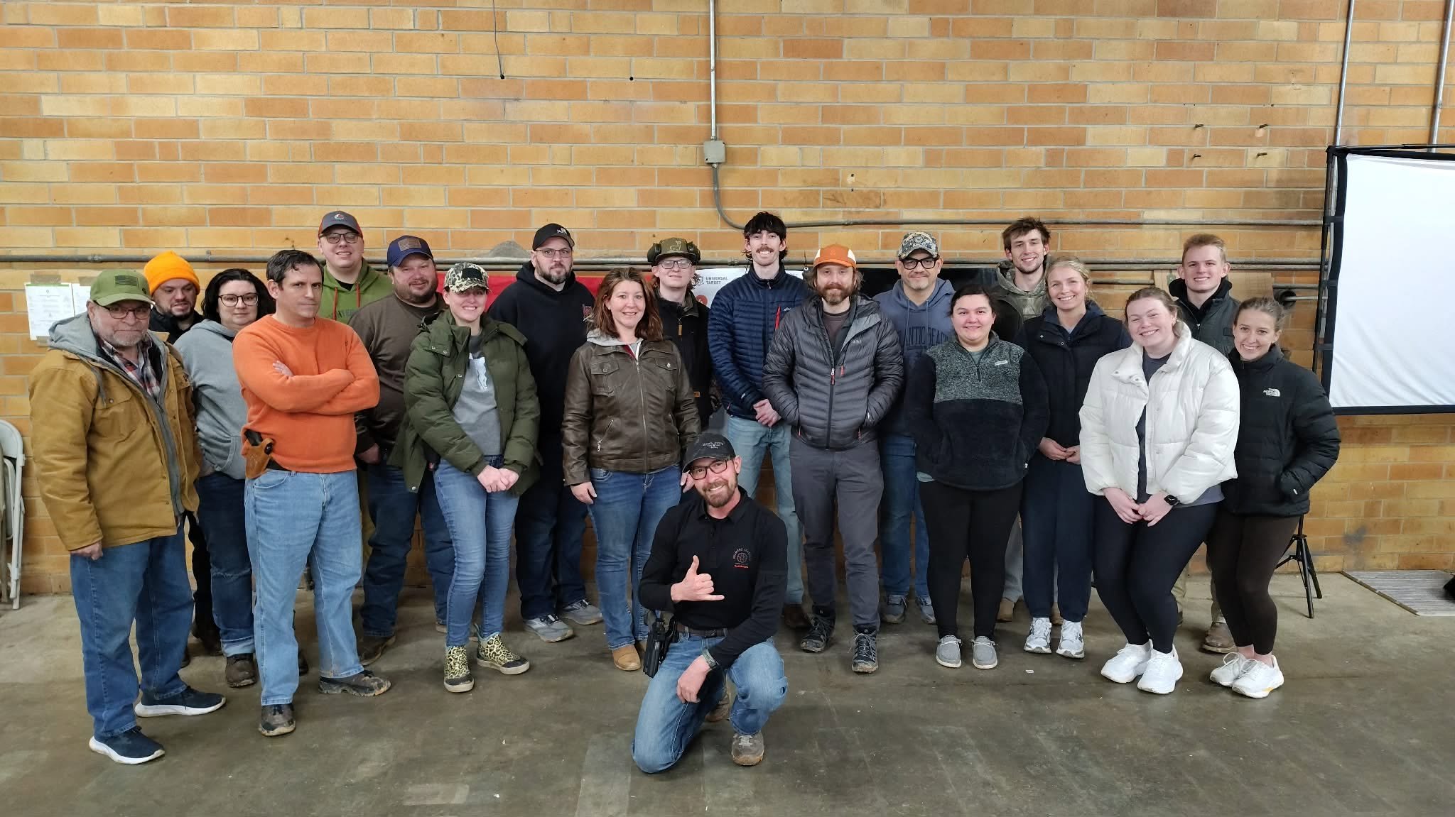 Group of approximately 20 people indoors in front of a brick wall, posing for a photo. They are dressed in casual and outdoor clothing, some wearing hats and glasses. One person is kneeling in the front, giving a thumbs up.