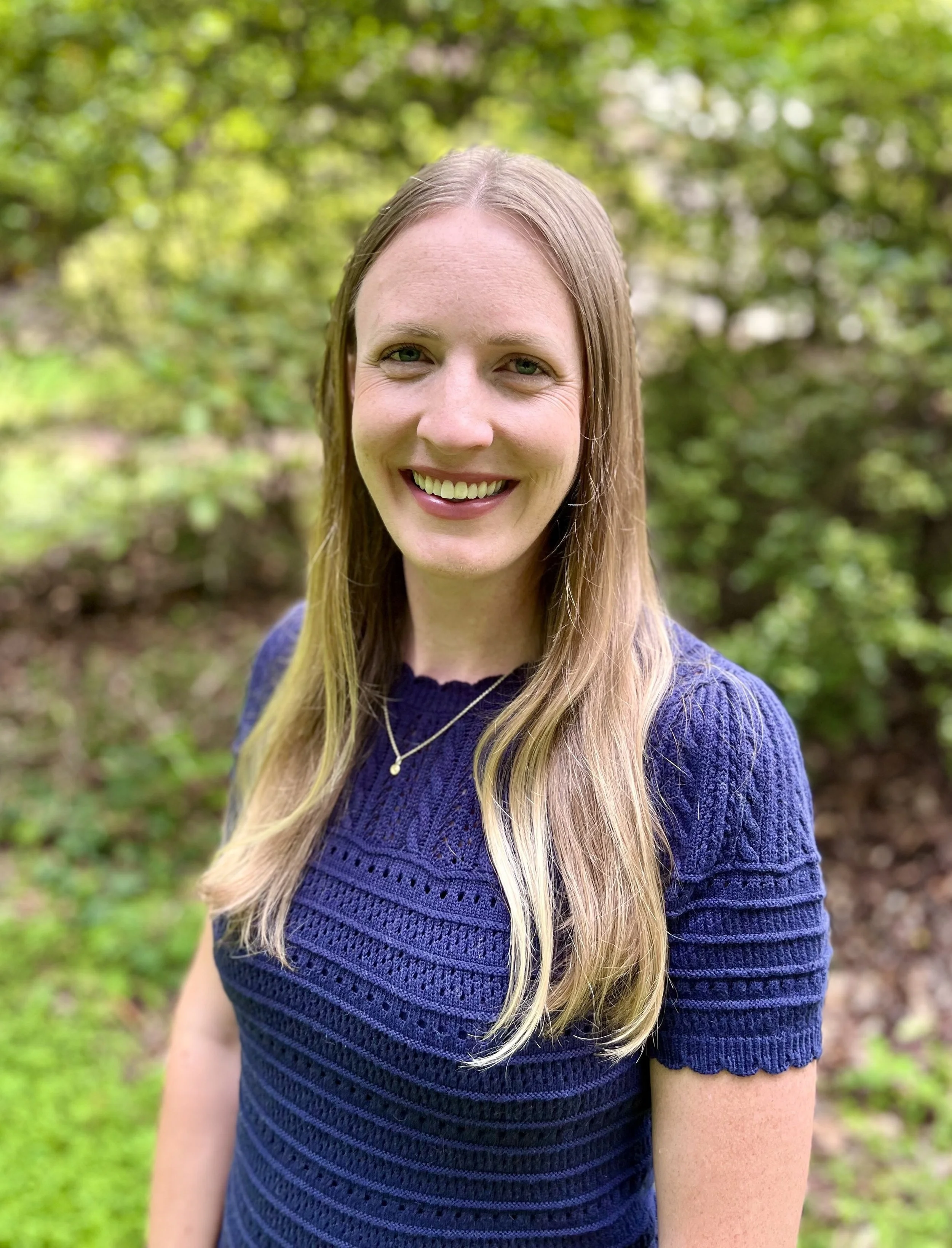 smiling myo functional therapist woman in blue shirt and blonde hair with a blurred greenery background