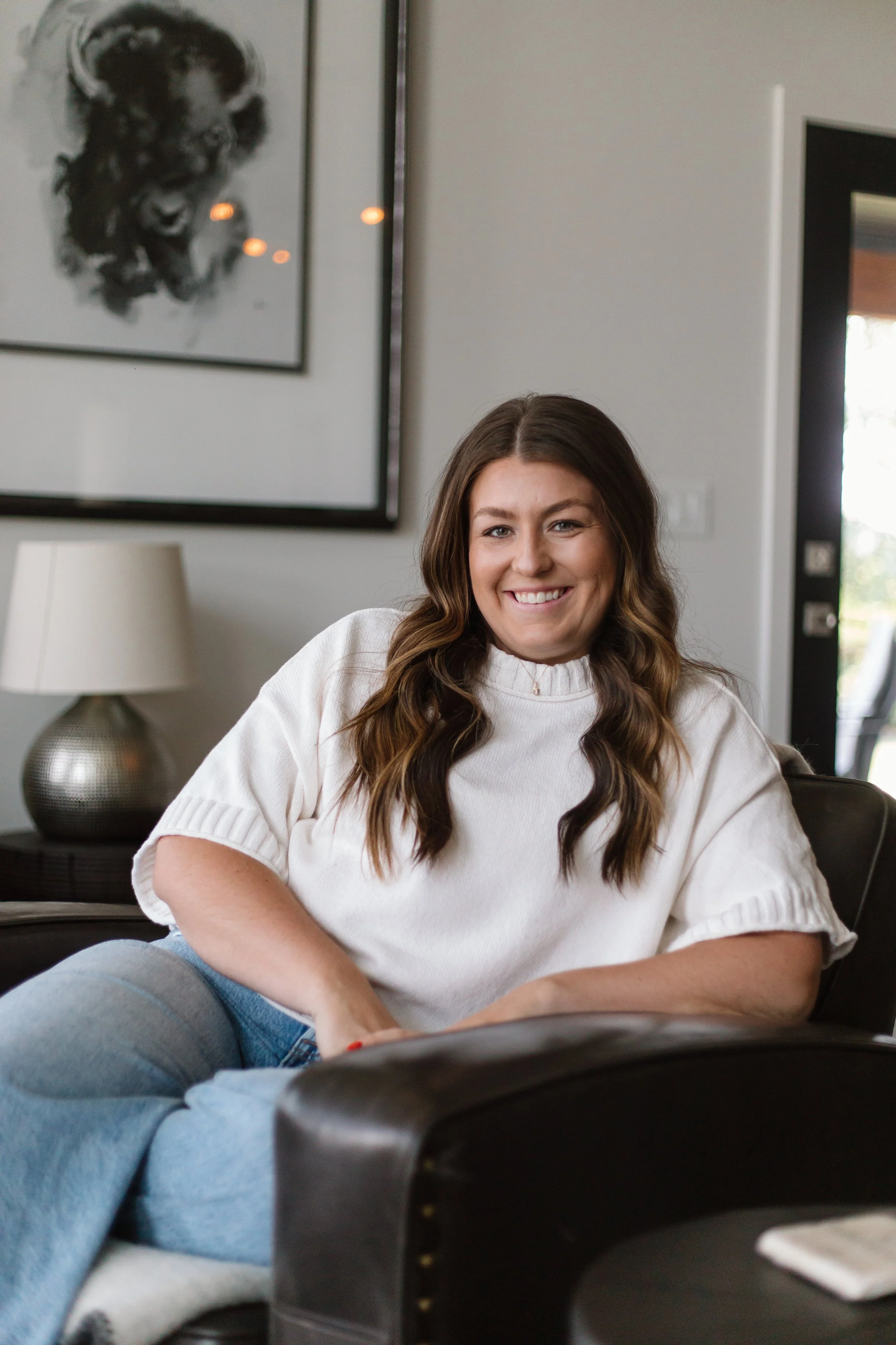 A woman with long brown hair, wearing a white sweater, smiling and sitting on a black sofa in a modern living room. There is a beige lamp on a side table and gray artwork on the wall behind her, with a door visible to the right.