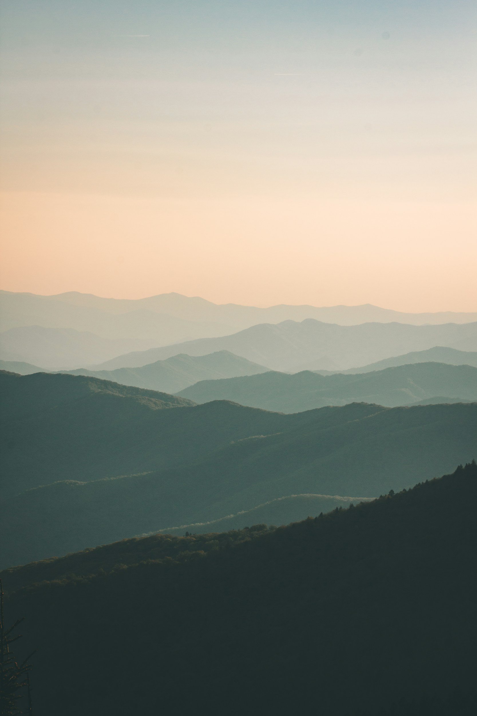 Hilly landscape with multiple layers of mountains during sunset or sunrise, with a soft pastel sky.